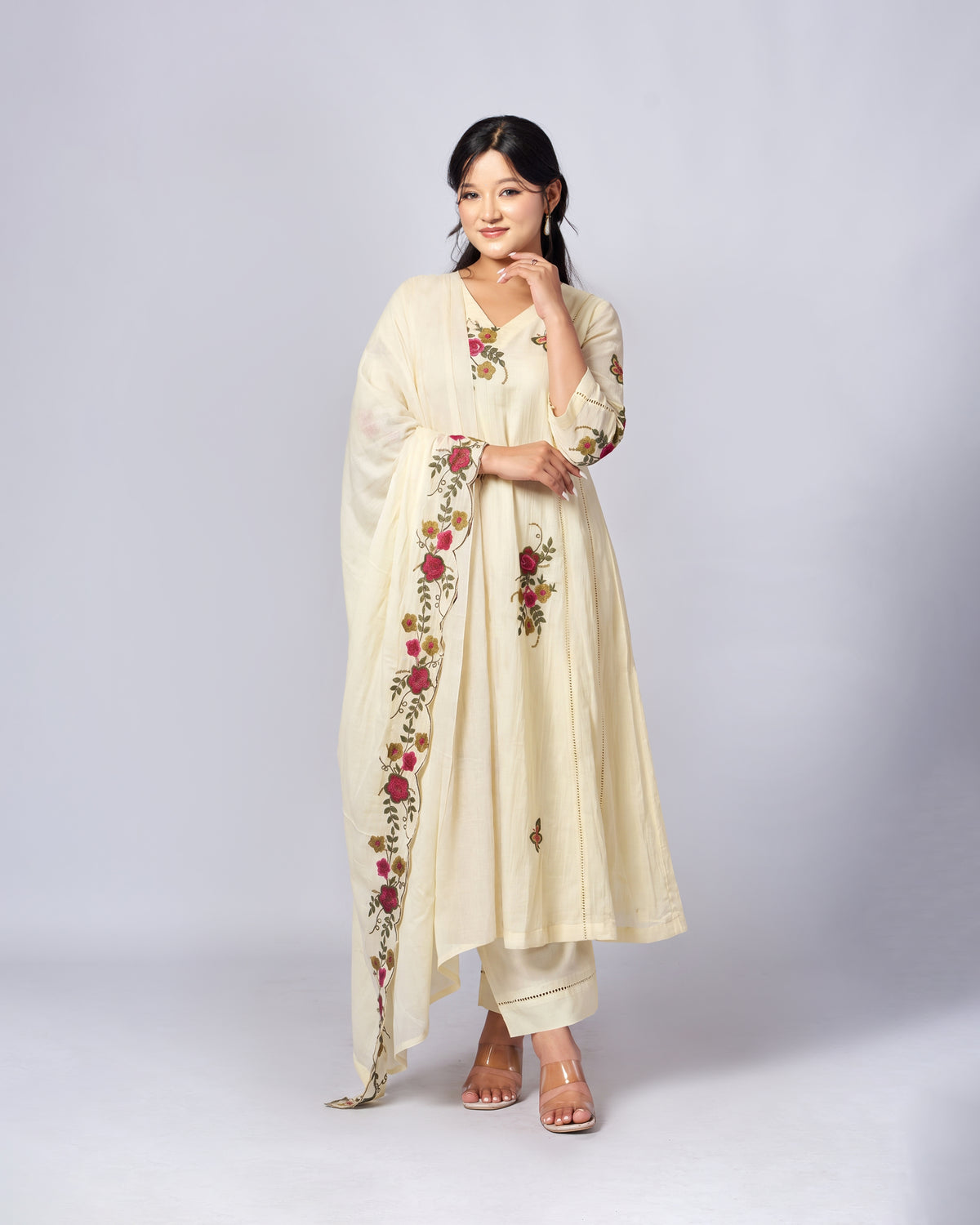 A woman modeling a cream-colored Indian ethnic suit set with floral embroidery. The set includes a long kurta, matching trousers, and a dupatta draped over her shoulder.