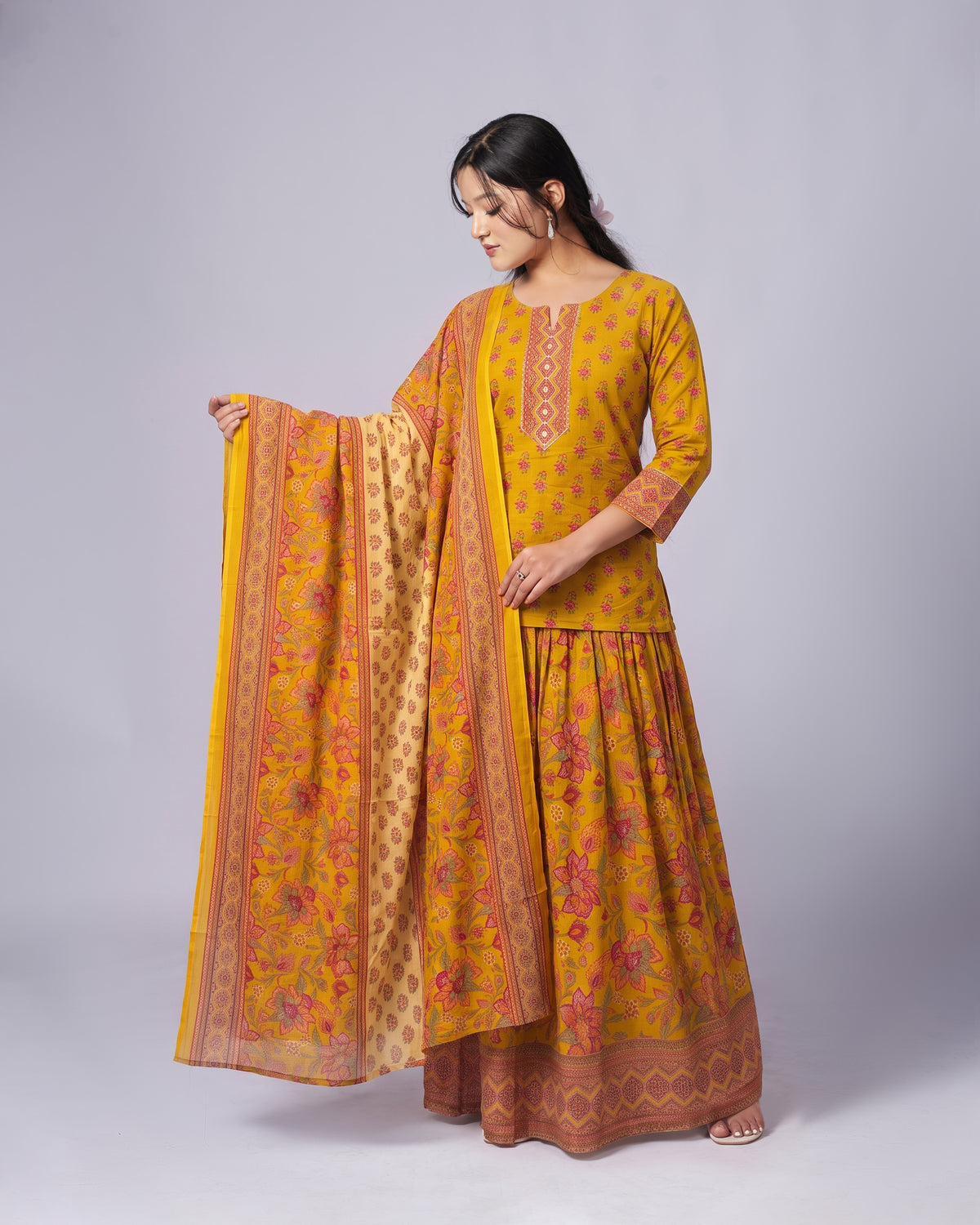 A woman modeling a traditional Indian outfit consisting of a mustard yellow kurta with floral prints, a matching flared skirt, and a coordinating dupatta, set against a plain light gray studio background.