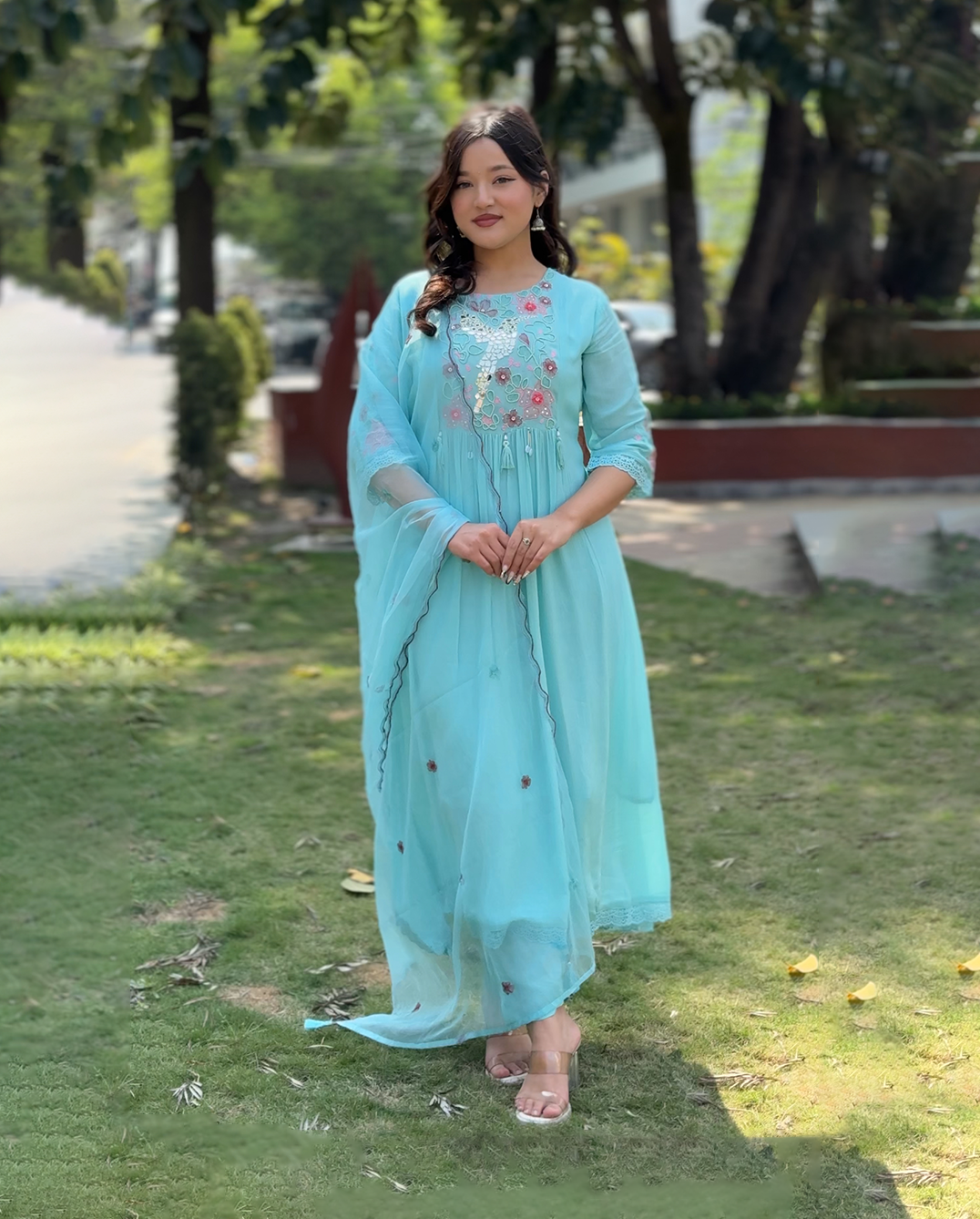 Woman in a light blue traditional outfit standing outdoors on grass with trees in the background