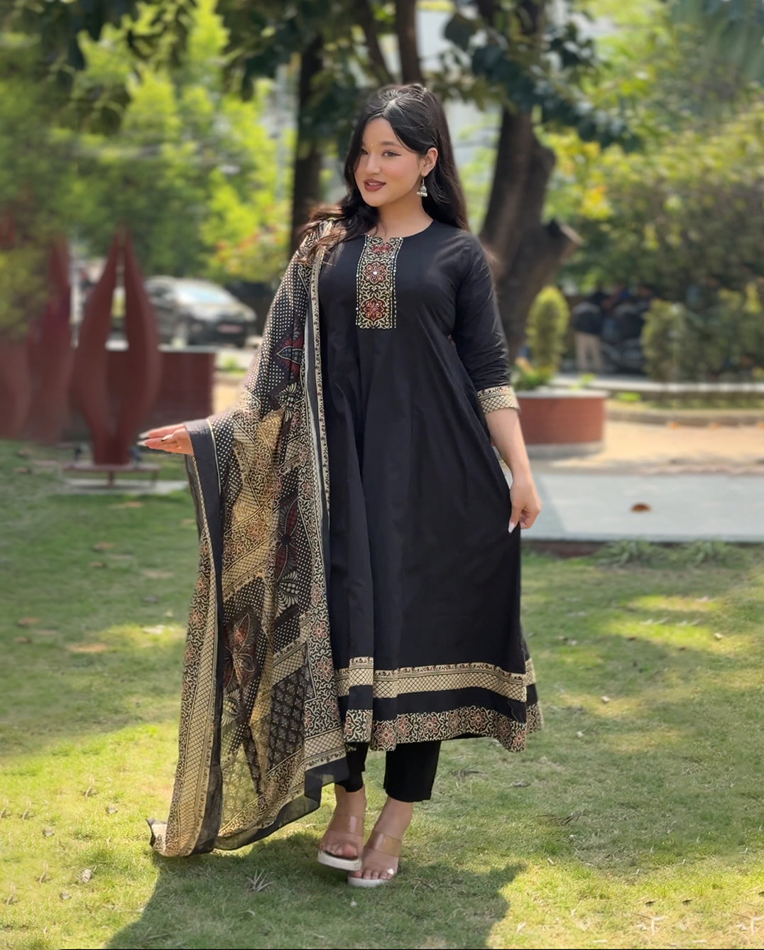 Woman in a black traditional outfit with a patterned shawl standing outdoors.