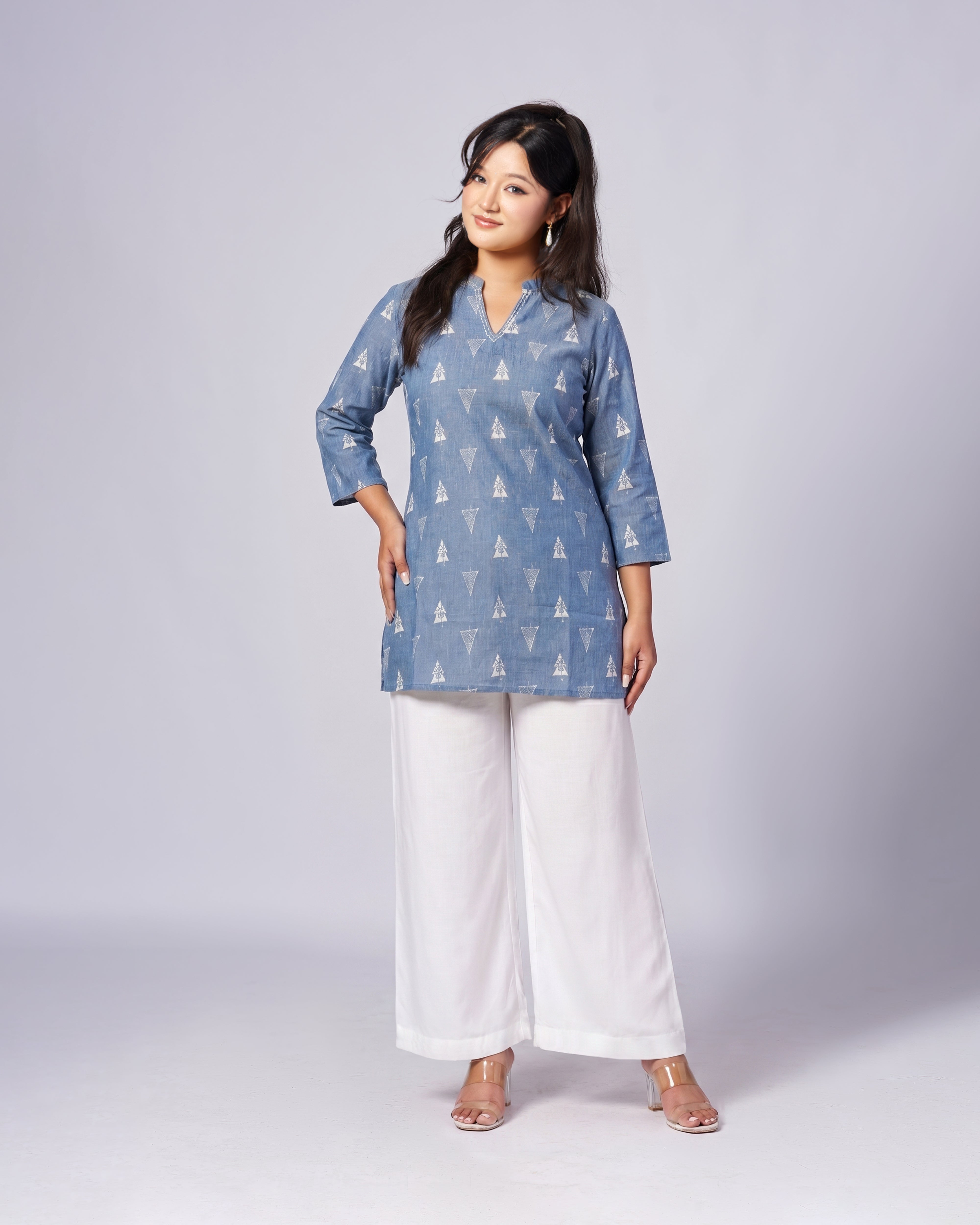 A woman modeling a light blue kurta with a white geometric pattern, paired with white wide-leg pants and clear high-heeled sandals, against a light gray studio background.