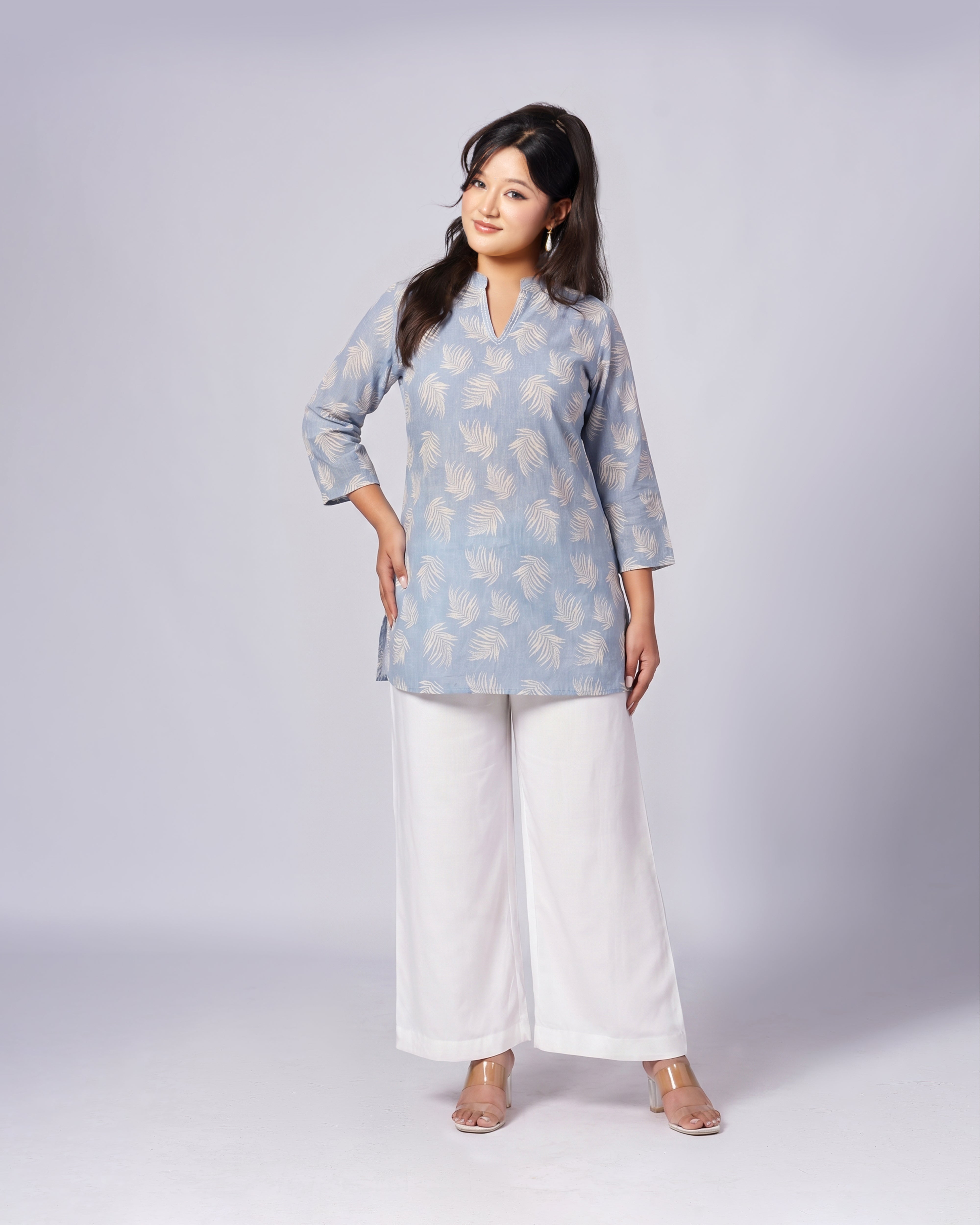 A woman modeling a light blue kurta with a beige palm leaf pattern, paired with white wide-leg pants, against a light gray studio background.
