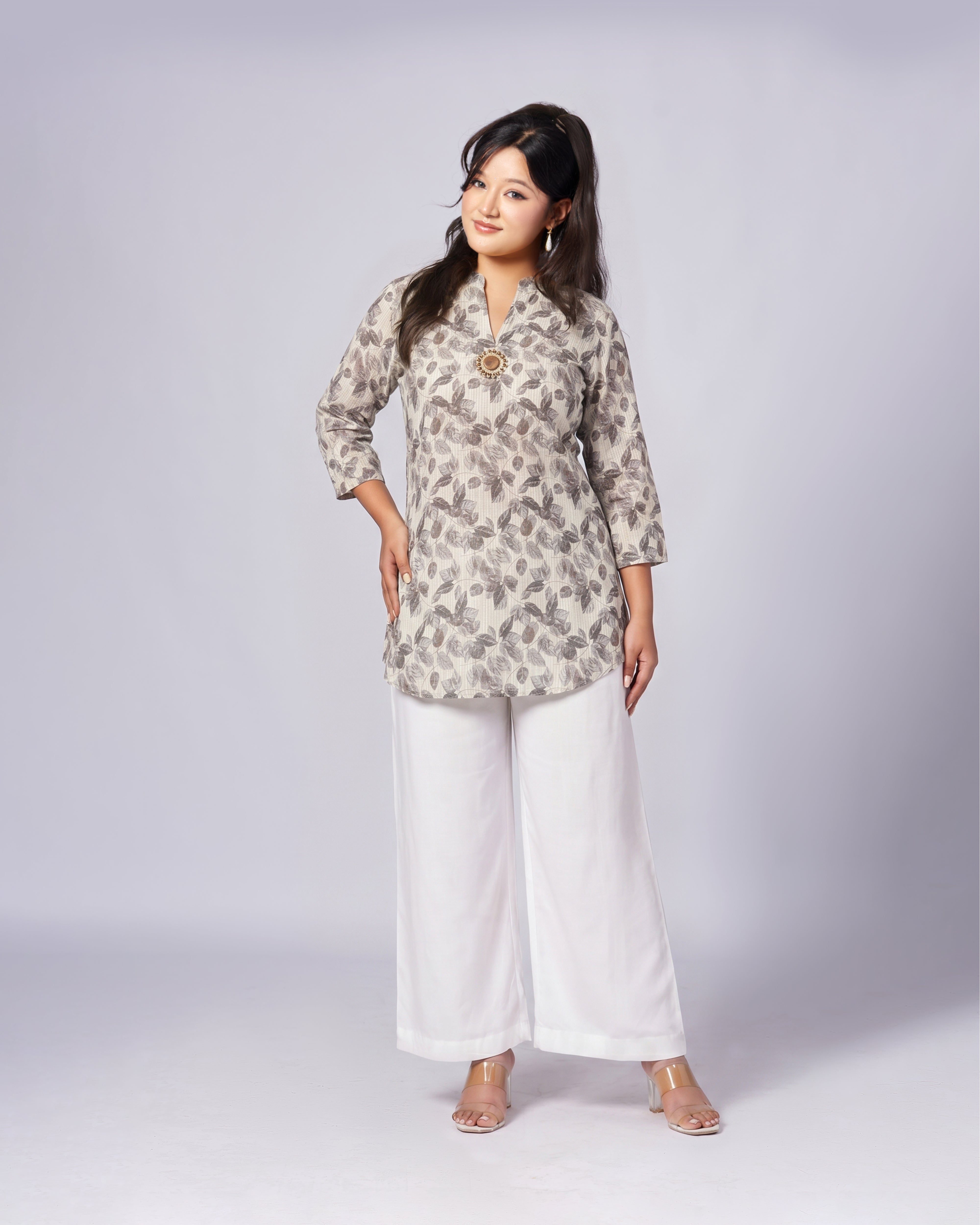 A woman modeling a three-quarter sleeve kurta with a gray and beige abstract pattern, paired with white wide-leg pants, standing against a light gray studio background.