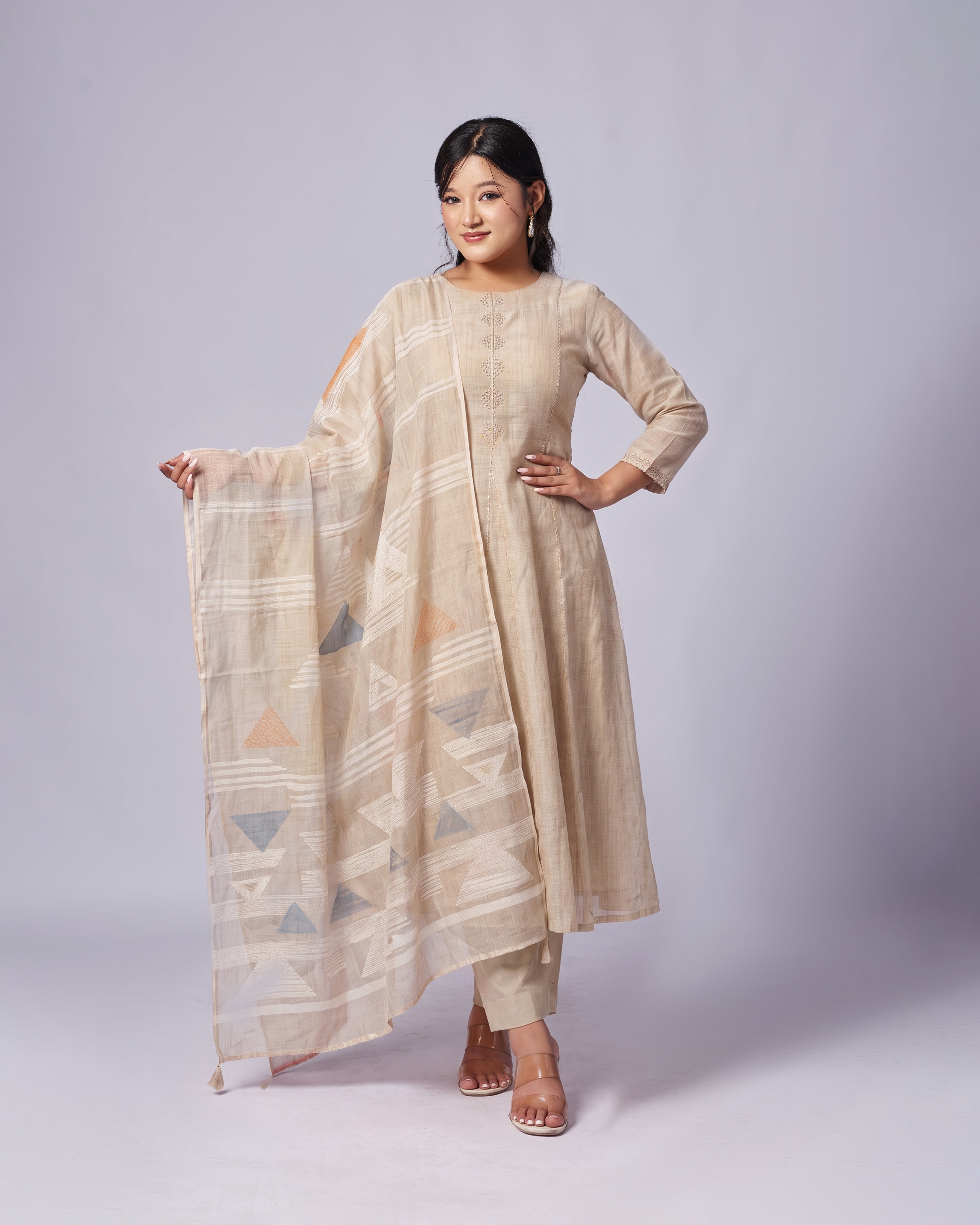 A woman modeling a beige three-piece traditional outfit consisting of a long-sleeve kurta, matching trousers, and a patterned dupatta with geometric triangle designs, set against a plain light gray studio background.
