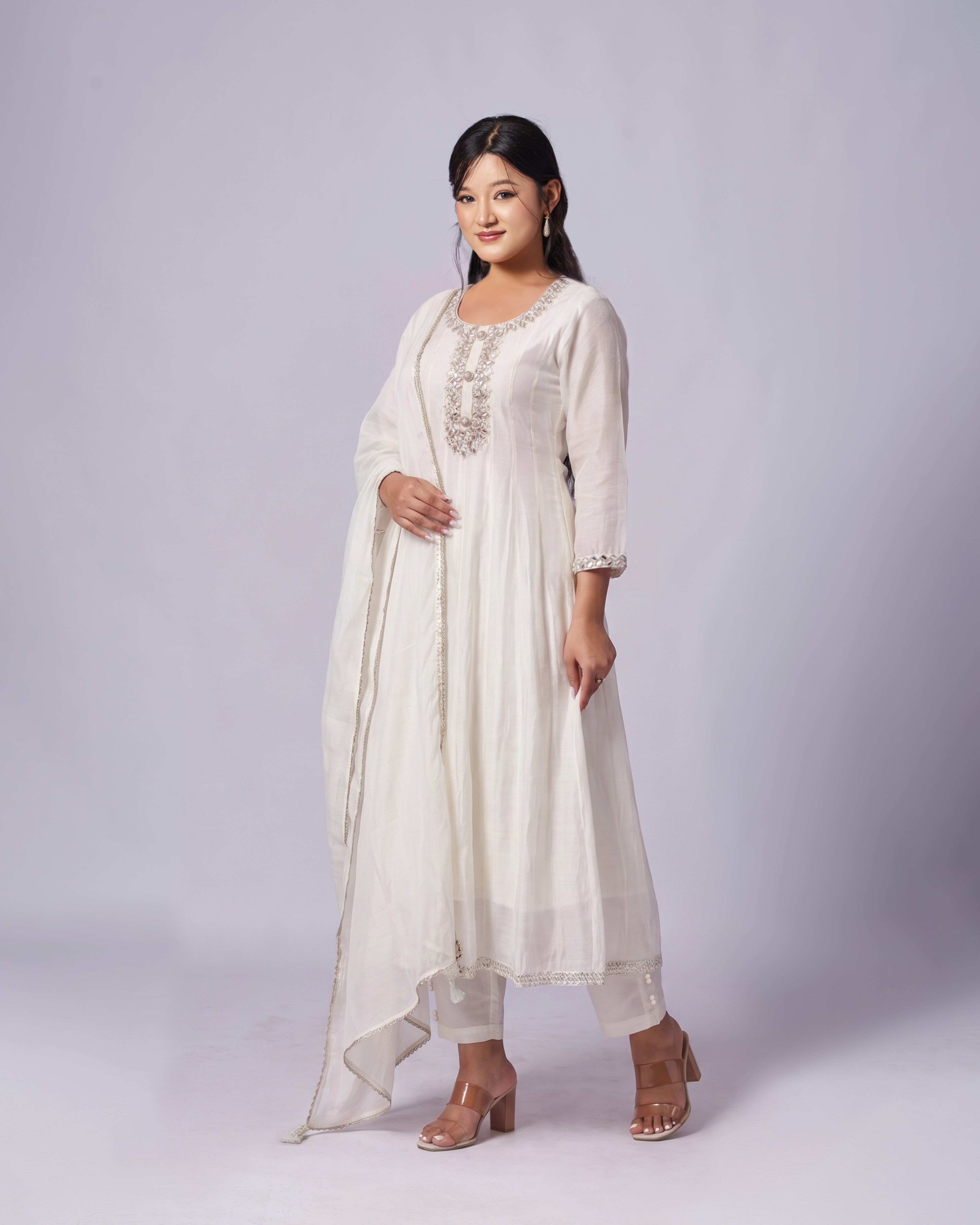 A woman modeling a traditional white Indian salwar kameez suit with a matching dupatta, featuring silver embroidery on the chest and sleeves, set against a plain light gray studio background.