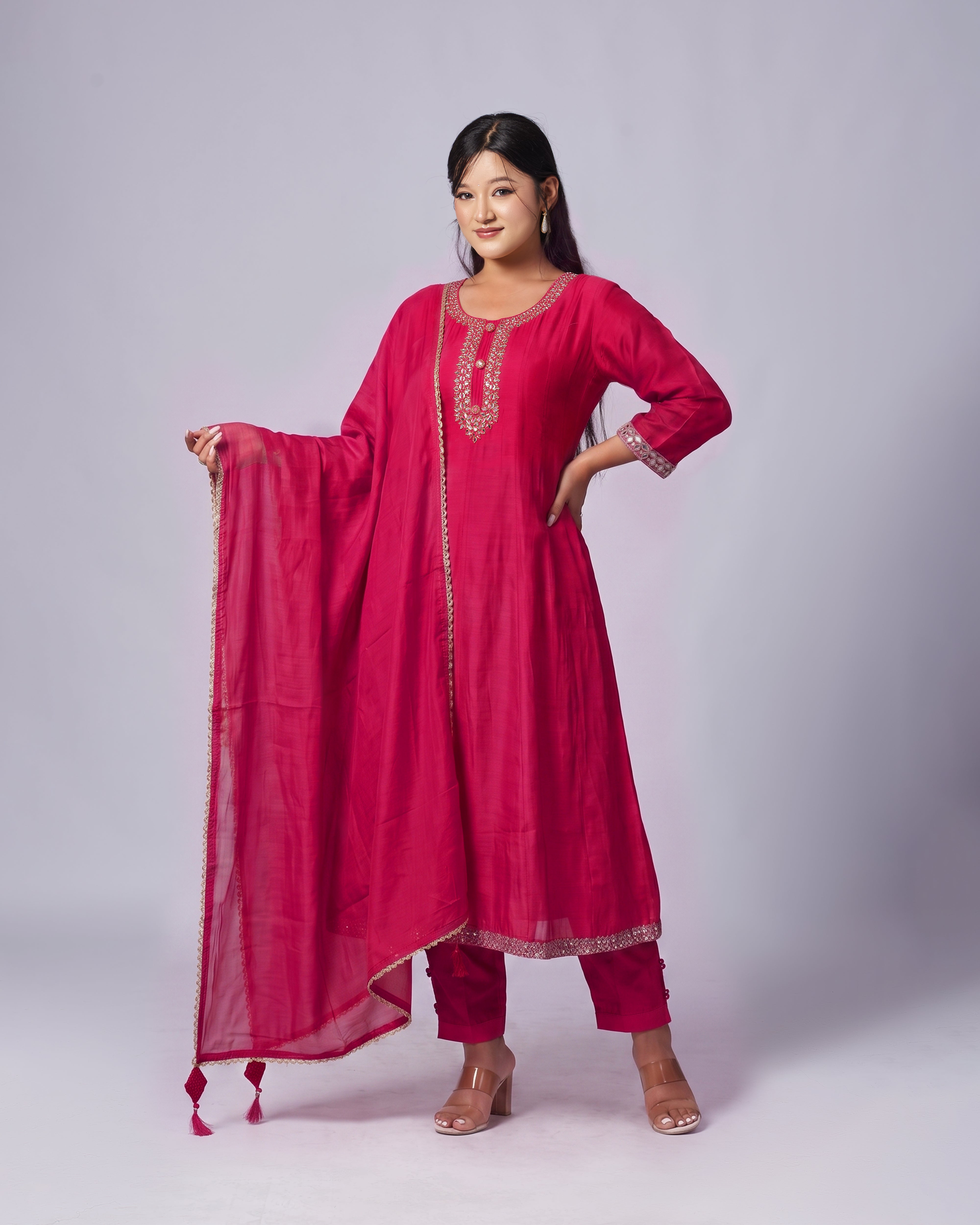 A woman modeling a bright pinkish-red Indian ethnic salwar kameez suit with a matching dupatta, featuring gold embroidery on the neckline, sleeve cuffs, and hem, set against a plain light gray studio background.
