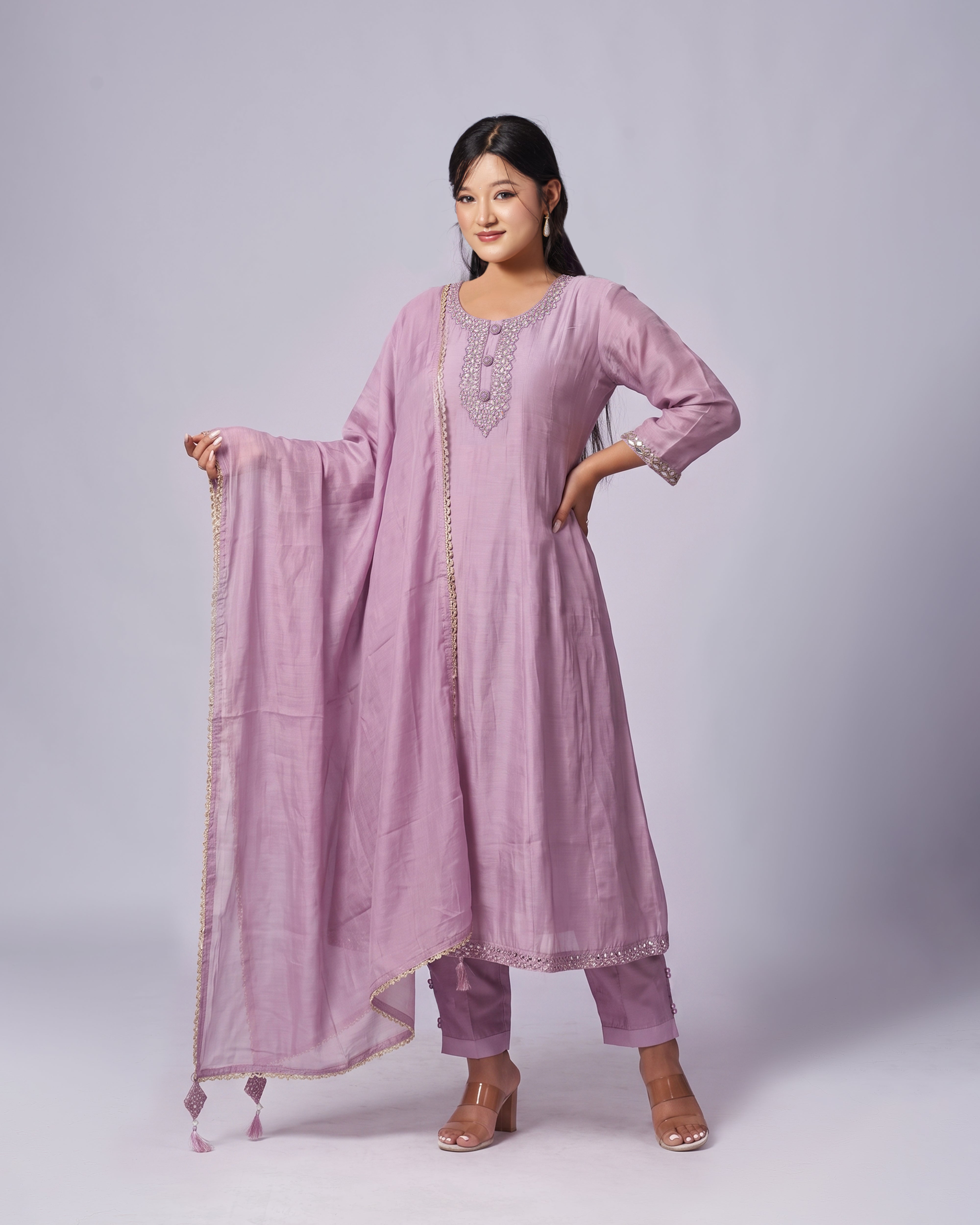 A woman modeling a light purple/lavender traditional Indian salwar kameez suit with matching pants and a dupatta, featuring silver embroidery on the neckline and borders, set against a plain light gray studio background.