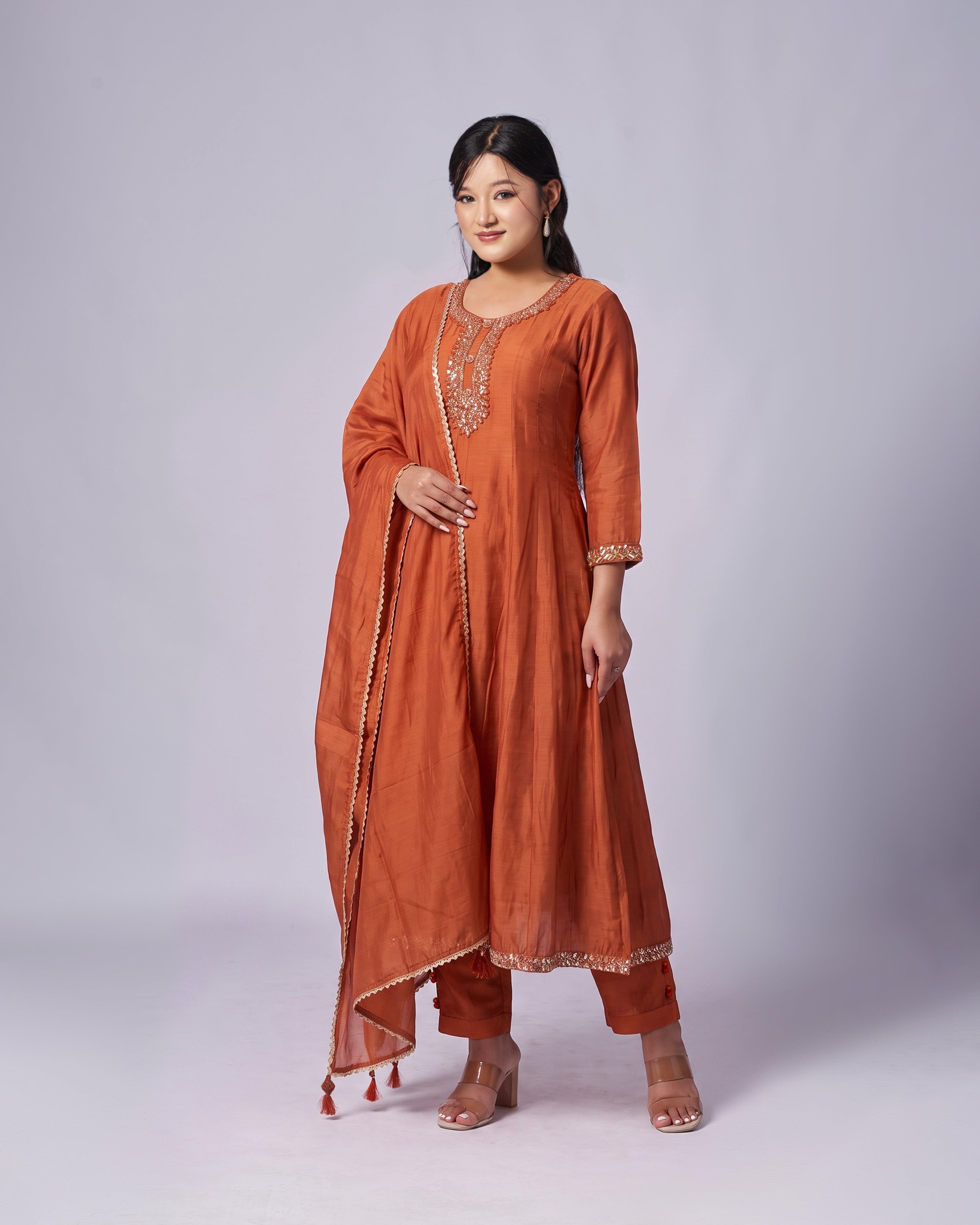 A woman modeling a rust-orange traditional Indian salwar kameez suit with a matching dupatta, featuring gold embroidery on the neckline and sleeve cuffs, set against a plain light gray studio background.
