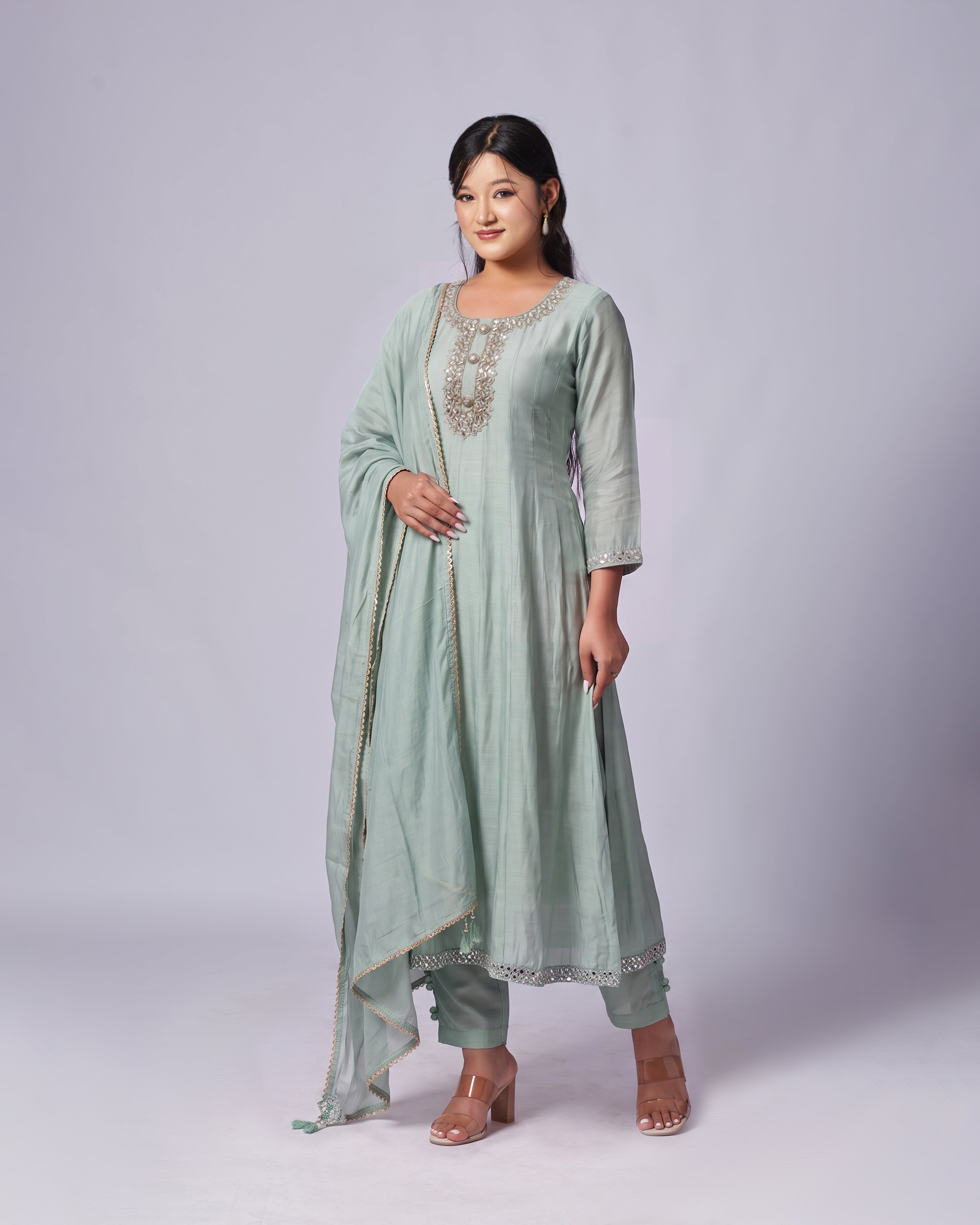 A woman modeling a light sage green traditional Indian outfit, featuring a straight kurta with silver embroidery at the neckline, matching pants, and a draped dupatta with gold border detailing, set against a plain light gray studio background.