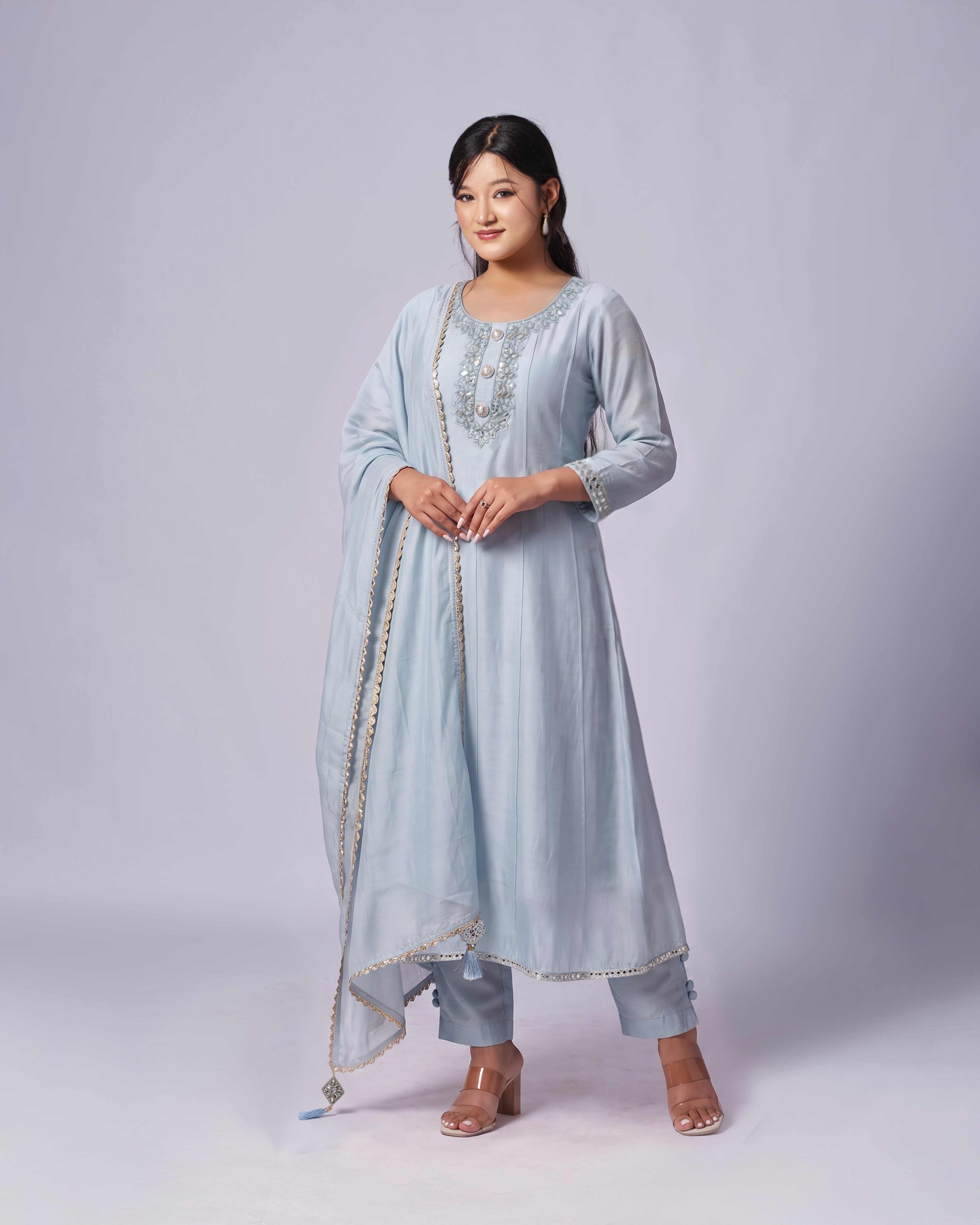A woman modeling a light blue traditional South Asian outfit with a matching dupatta, featuring silver embroidery on the neckline and gold zari detailing along the borders, set against a plain light gray studio background.
