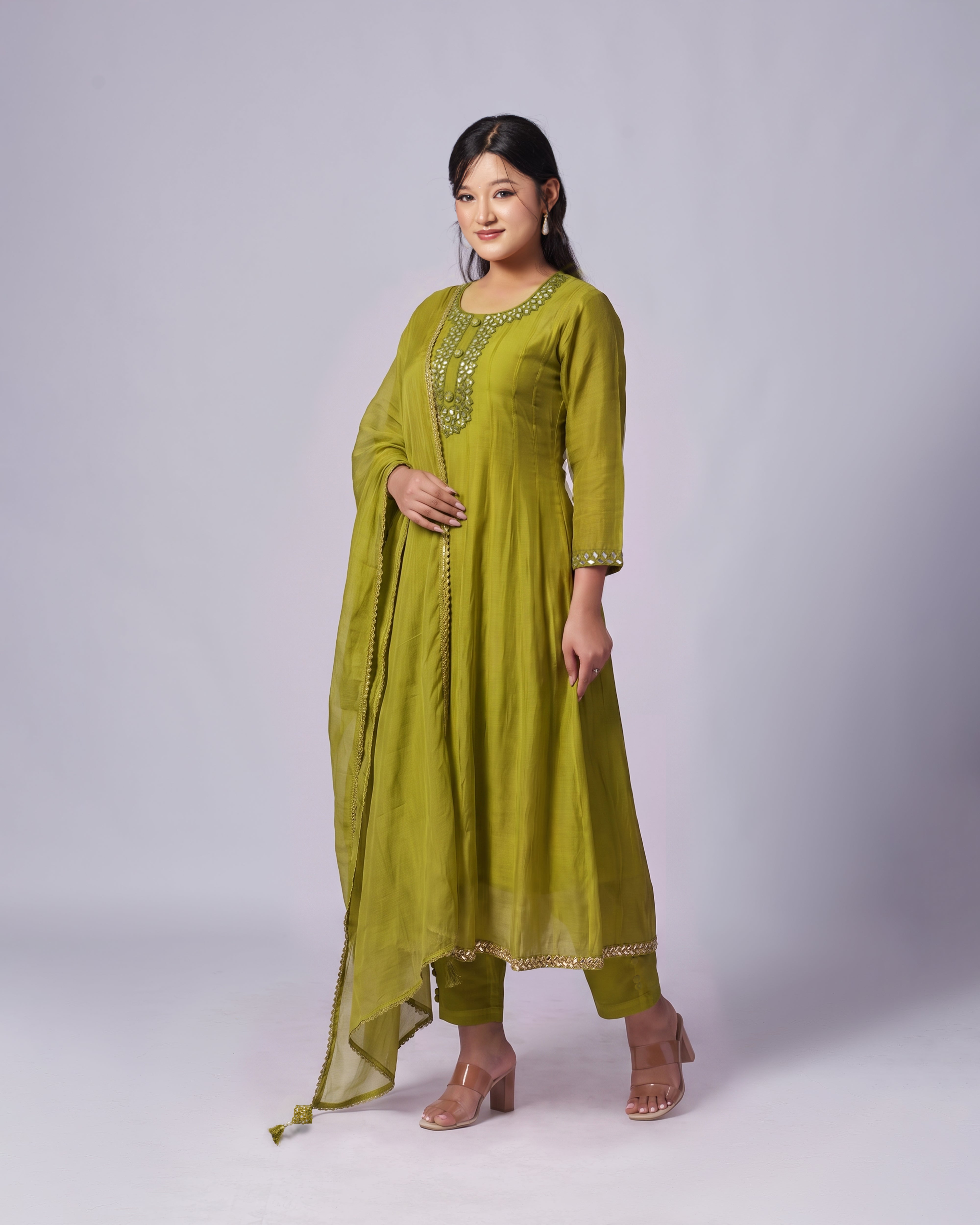 A woman modeling a three-piece olive green ethnic suit with gold embroidery on the neckline and hem, paired with matching pants and a draped dupatta, set against a plain light gray studio background.