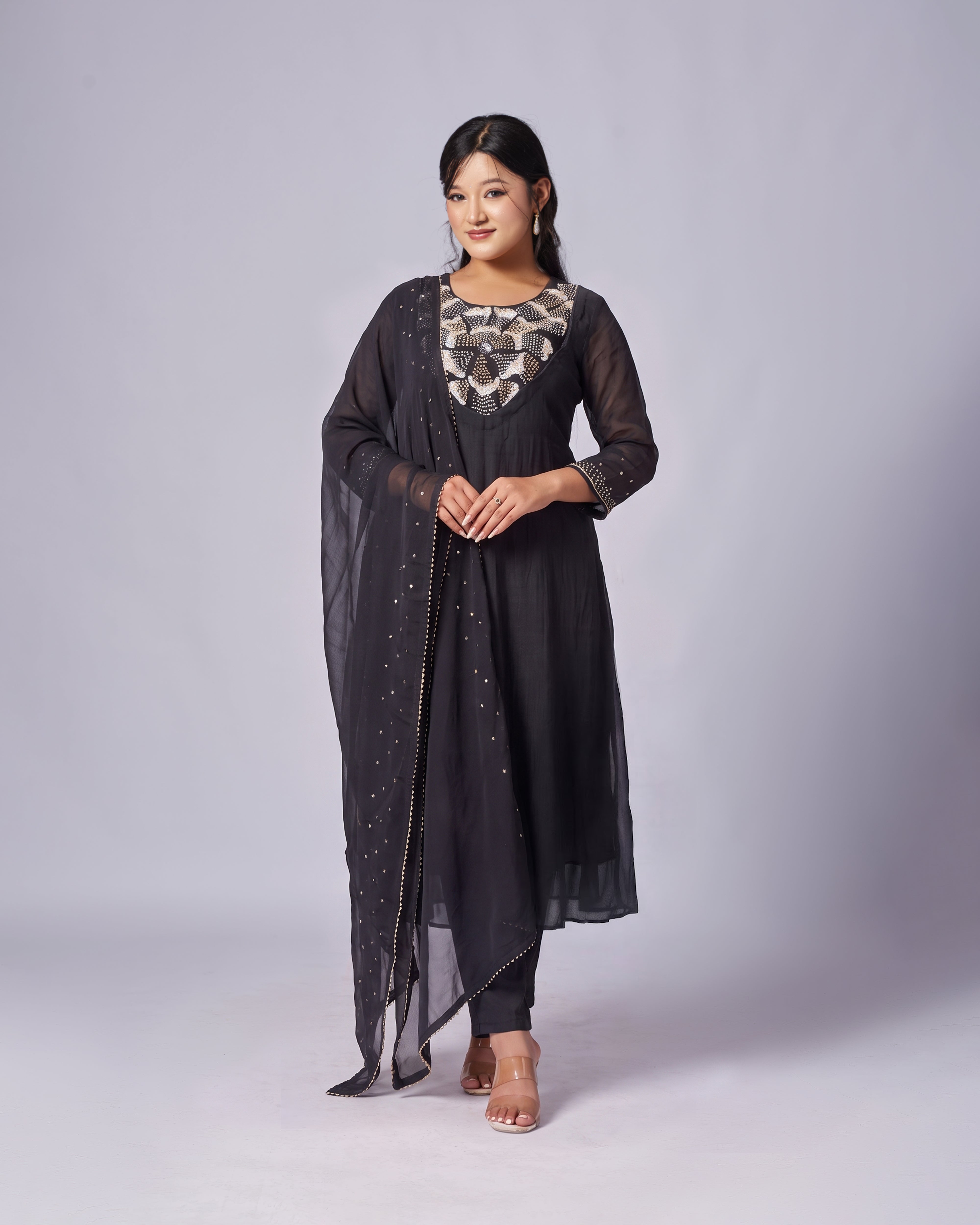 A woman modeling a black traditional outfit with a sheer embroidered dupatta, patterned neckline, and embellished skirt, photographed against a light gray studio background.