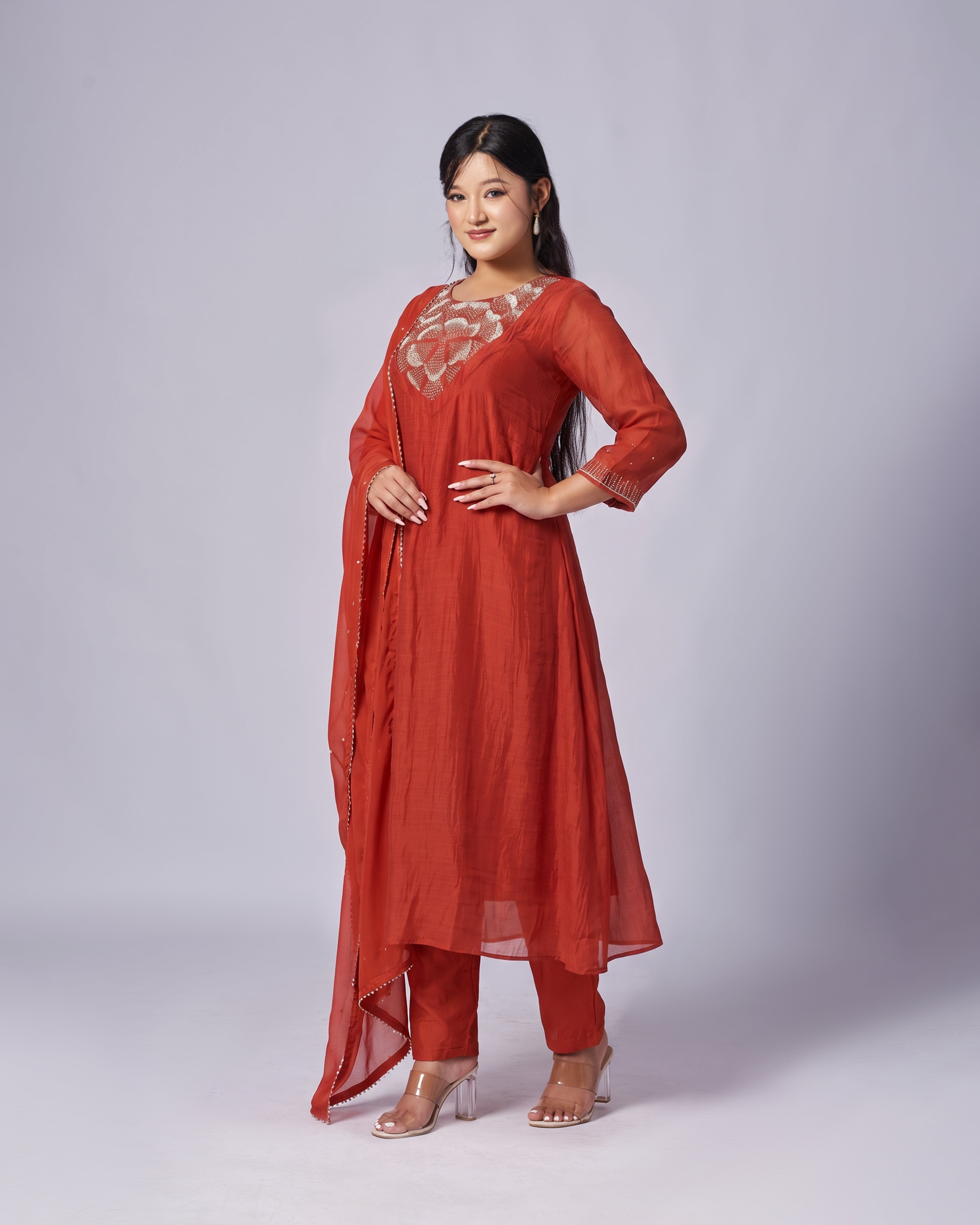 A woman modeling a rust-orange traditional Indian salwar kameez suit with a matching dupatta, featuring a sheer embroidered yoke and matching trousers, set against a light gray studio background.