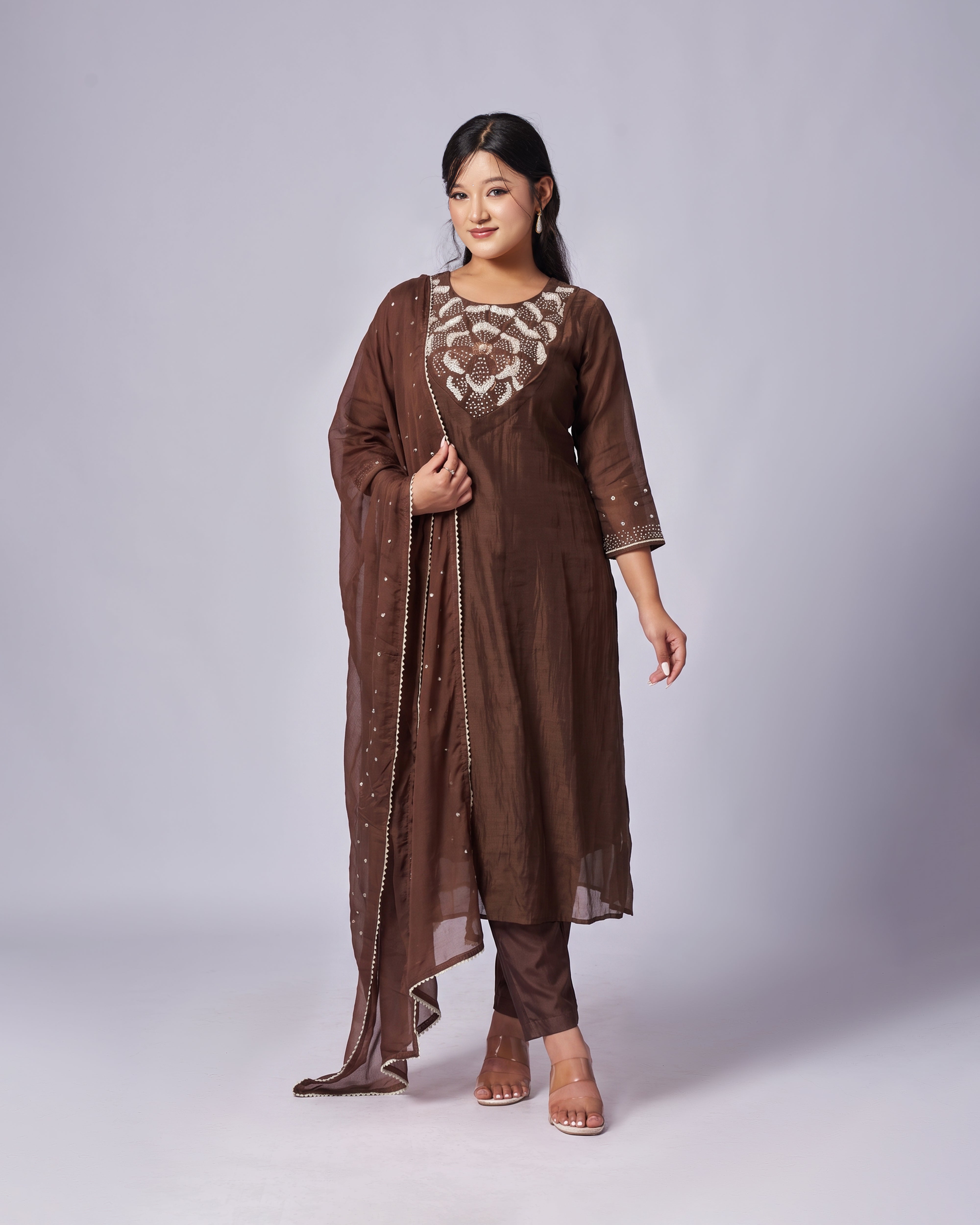 A woman modeling a brown traditional outfit with a matching sheer dupatta, featuring a printed floral neckline and embroidered details on the sleeves, set against a plain light gray studio background.