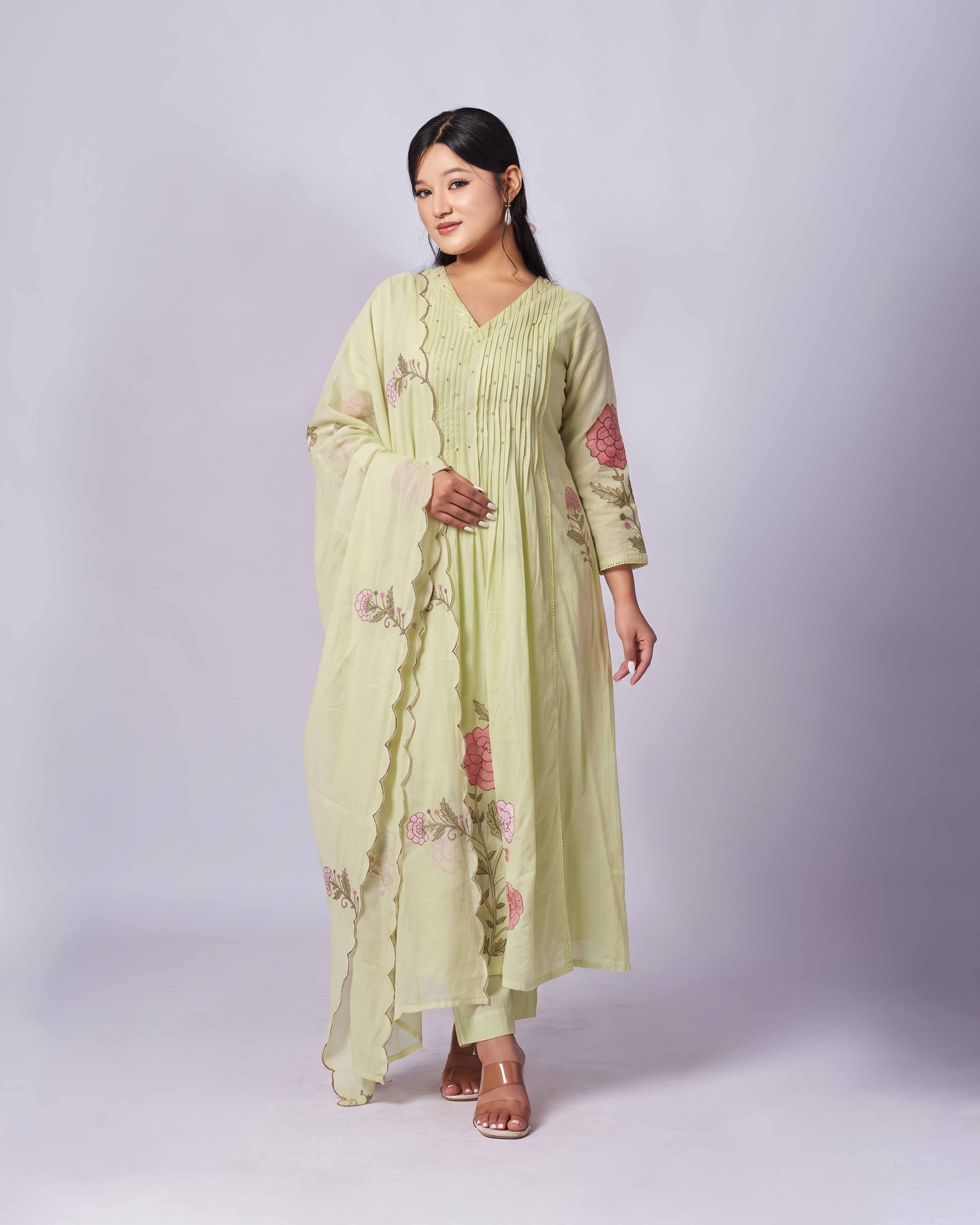 A woman modeling a light sage green kurta set with pink floral embroidery and a matching dupatta, standing against a light gray studio background.