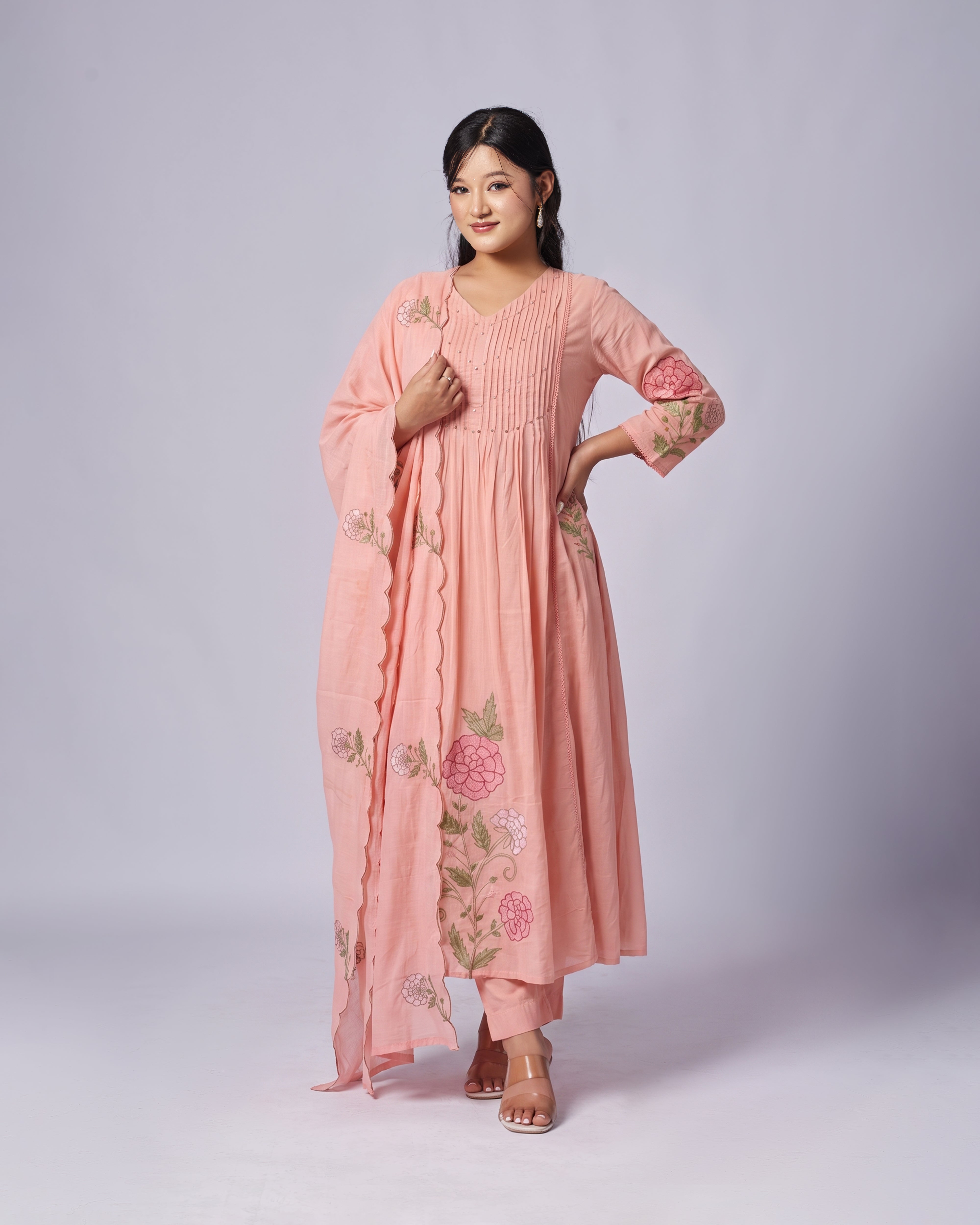 A woman modeling a peach-pink traditional kurta set with floral embroidery and a matching dupatta, photographed against a light gray studio background.