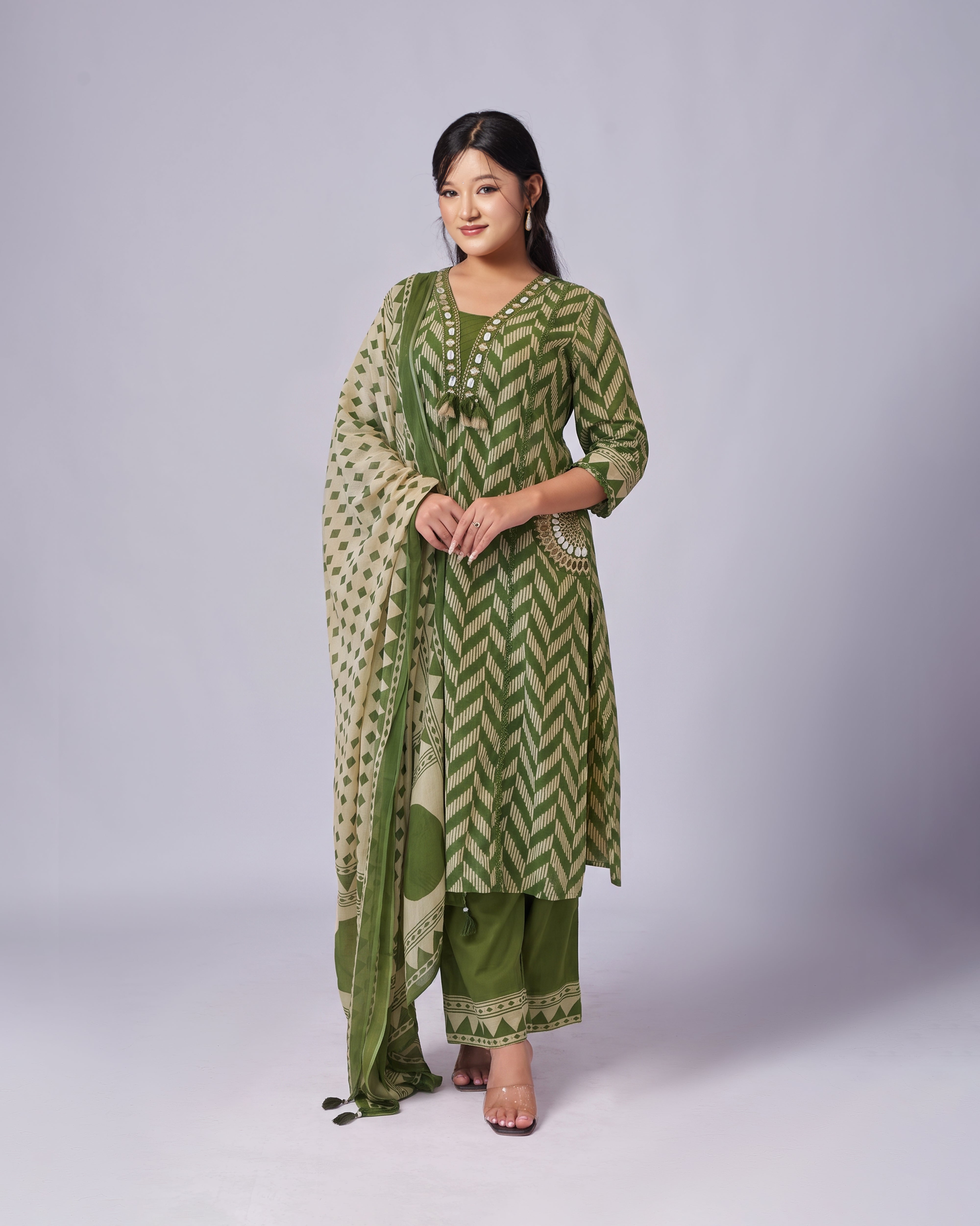A woman modeling a green and beige patterned traditional outfit with a matching dupatta, standing against a light gray studio background.