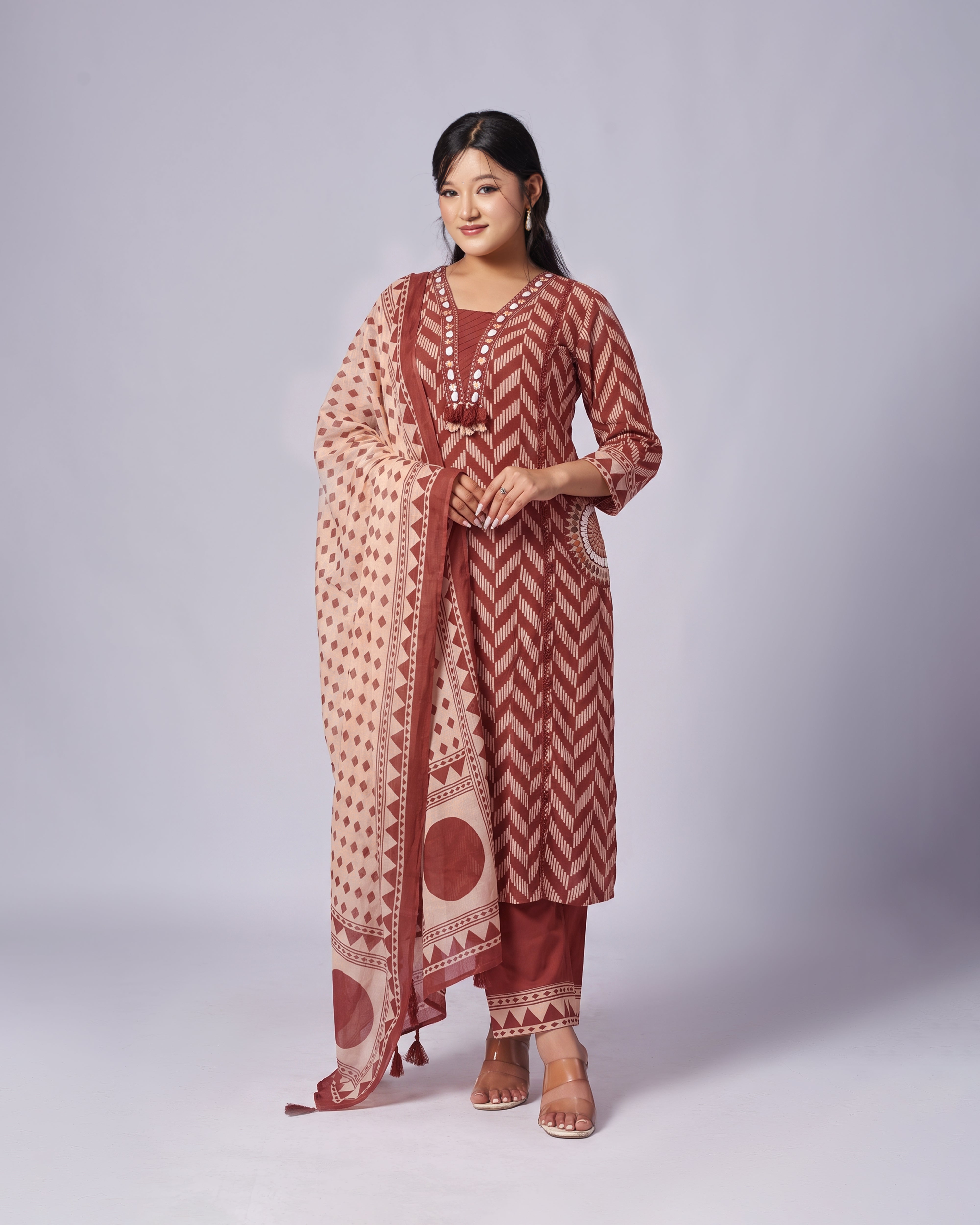 A woman modeling a traditional Indian salwar kameez suit set in rust red and beige. The set includes a rust red long-sleeve kurta with a zigzag pattern, matching pants, and a beige dupatta with red geometric patterns.