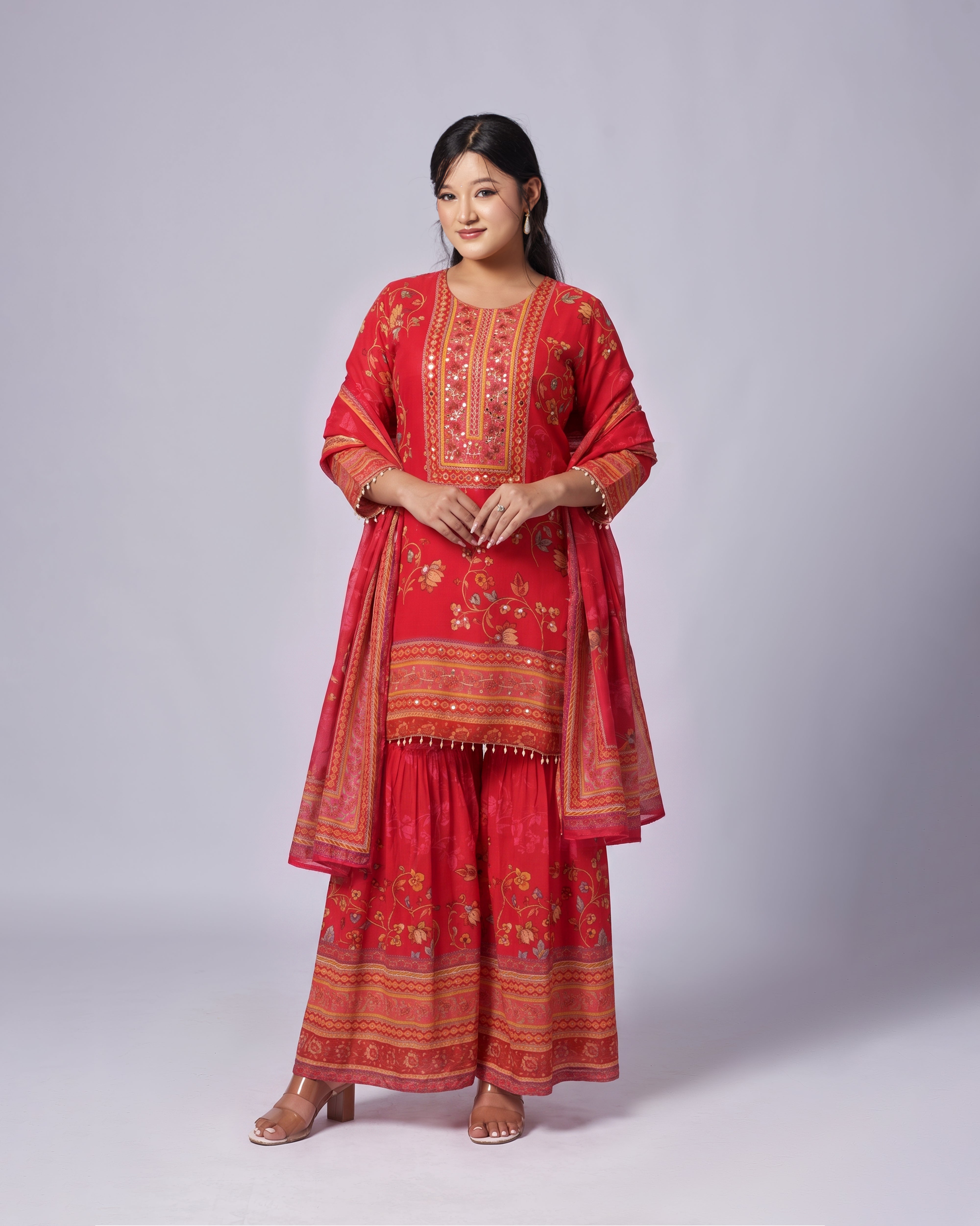 A woman modeling a traditional red Indian salwar kameez suit with intricate gold and multicolor embroidery, paired with matching wide-leg pants and a dupatta, set against a plain light gray studio background.