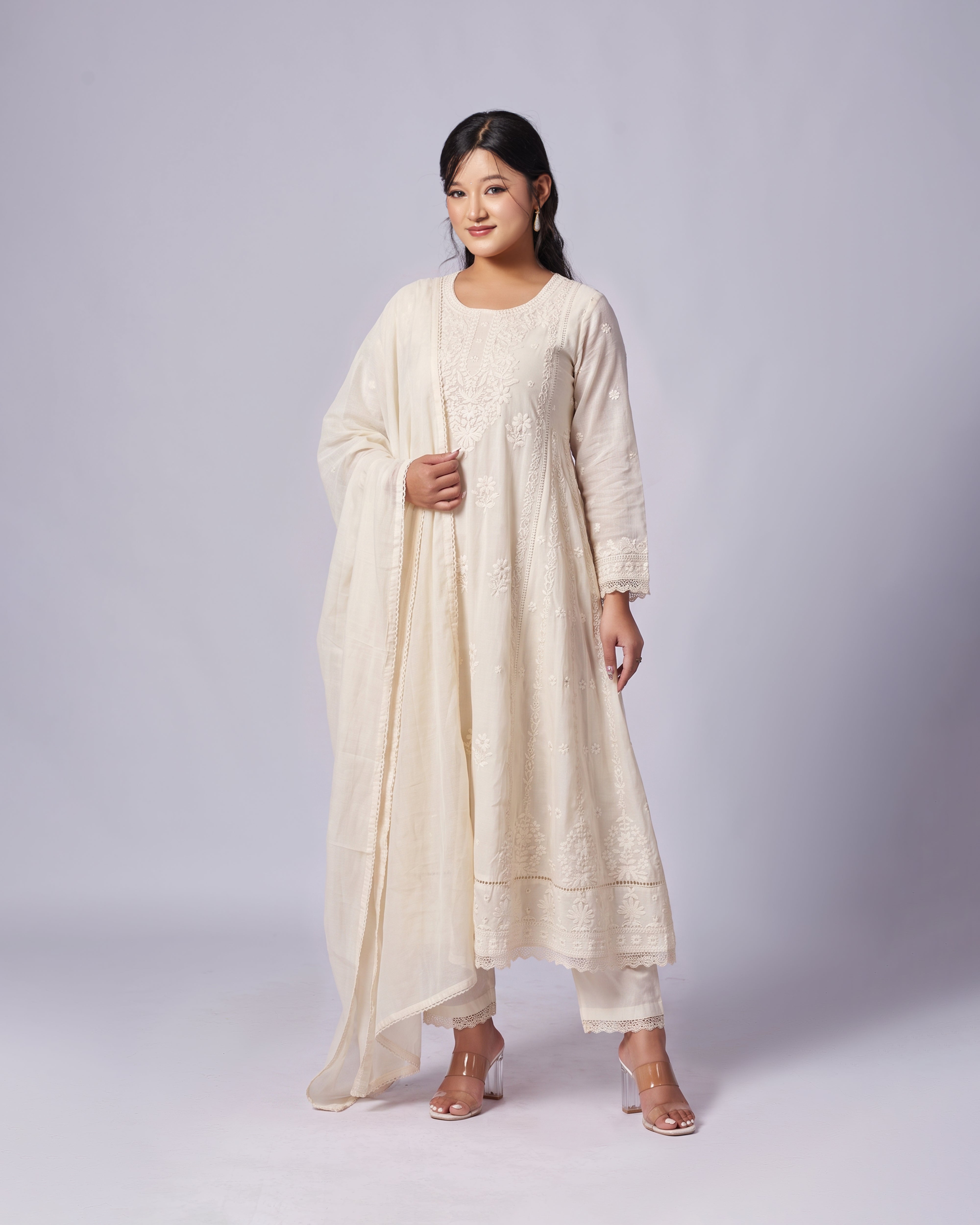 A woman modeling a cream-colored traditional South Asian outfit, featuring a long kurta with delicate embroidery, matching trousers, and a draped dupatta, set against a plain light gray studio background.