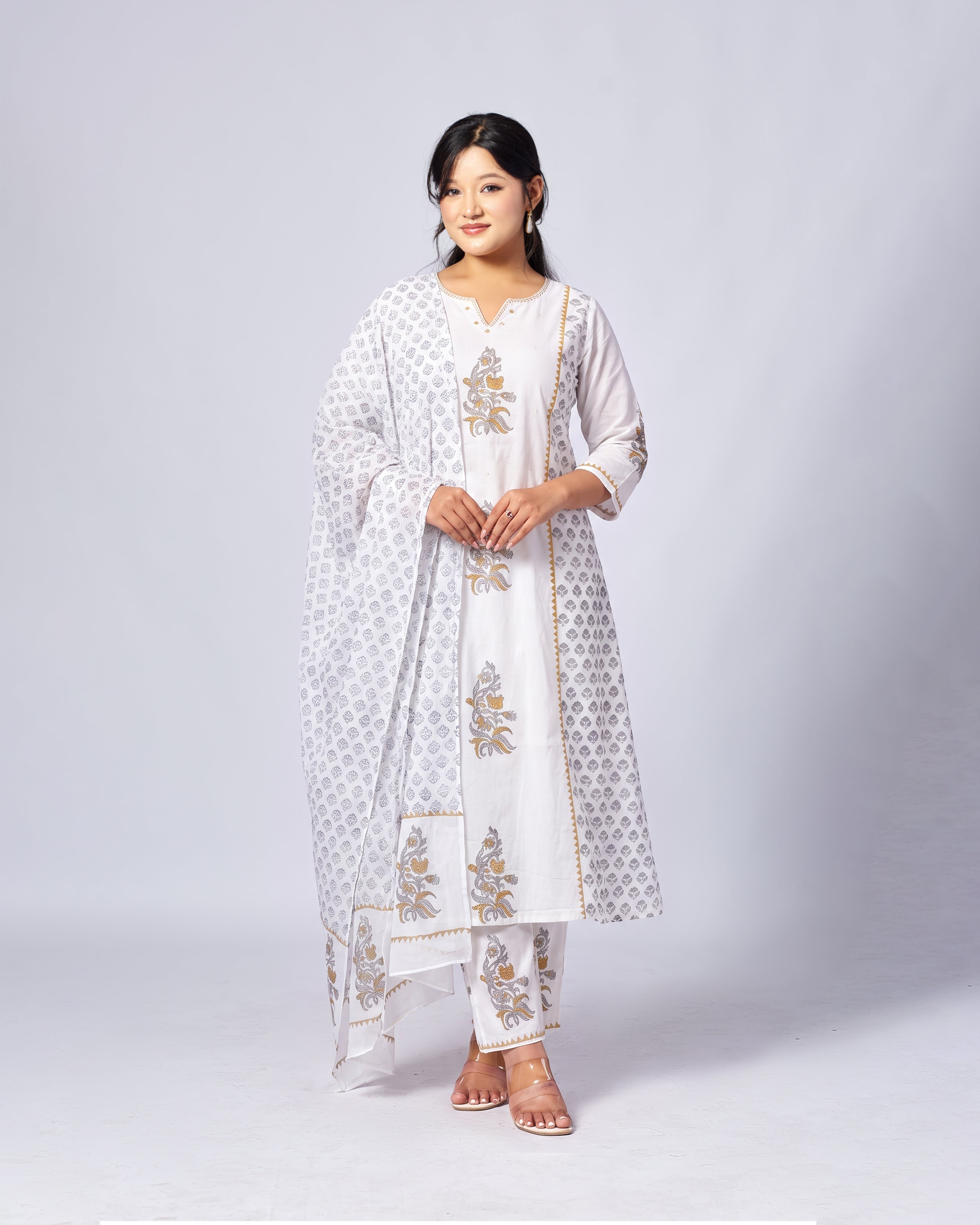 A woman modeling a white traditional South Asian outfit with gold floral embroidery and a matching white patterned dupatta, set against a light gray studio background.