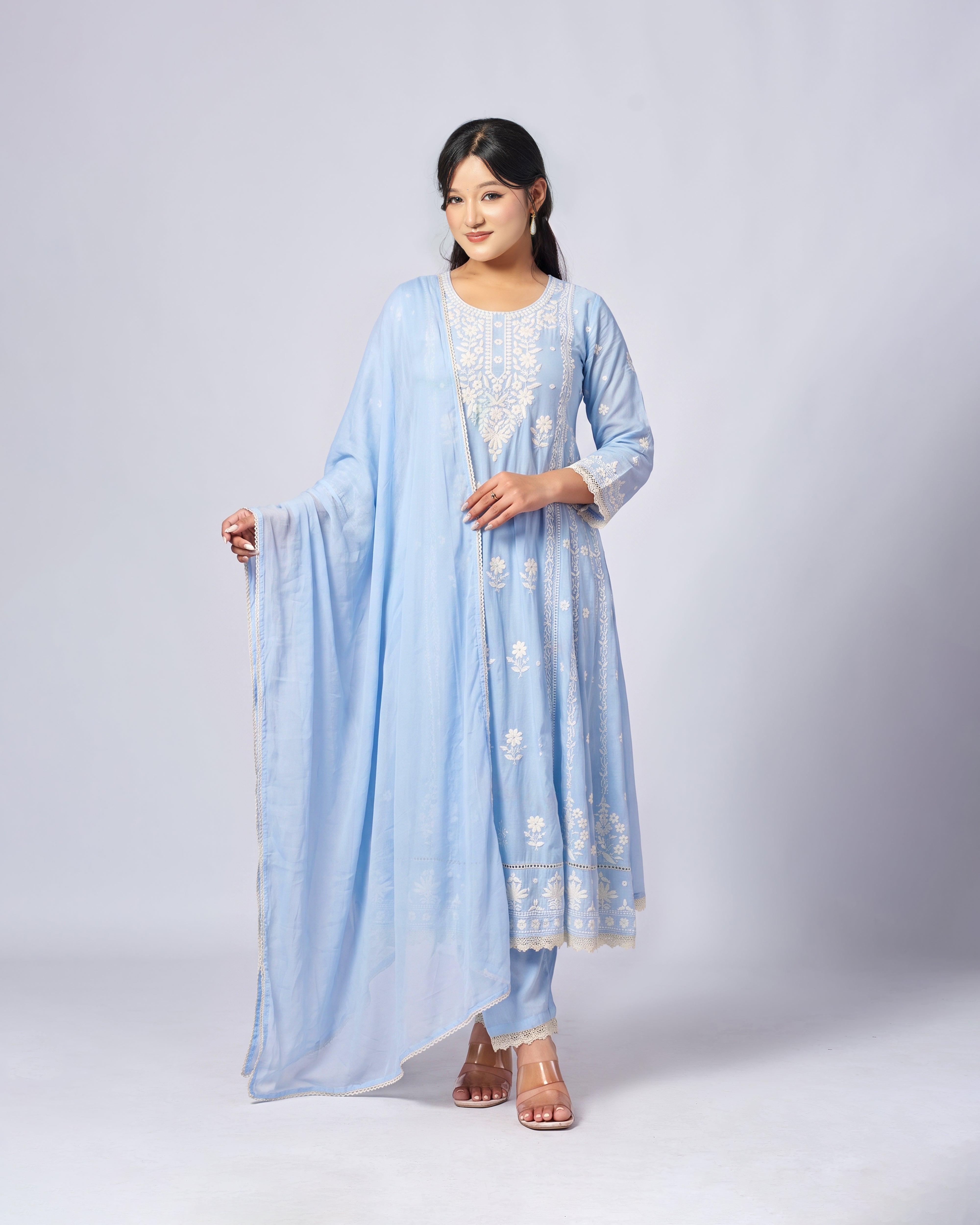 A woman modeling a light blue traditional outfit with white embroidery, featuring a long kurta, matching pants, and a draped dupatta, set against a plain light gray studio background.