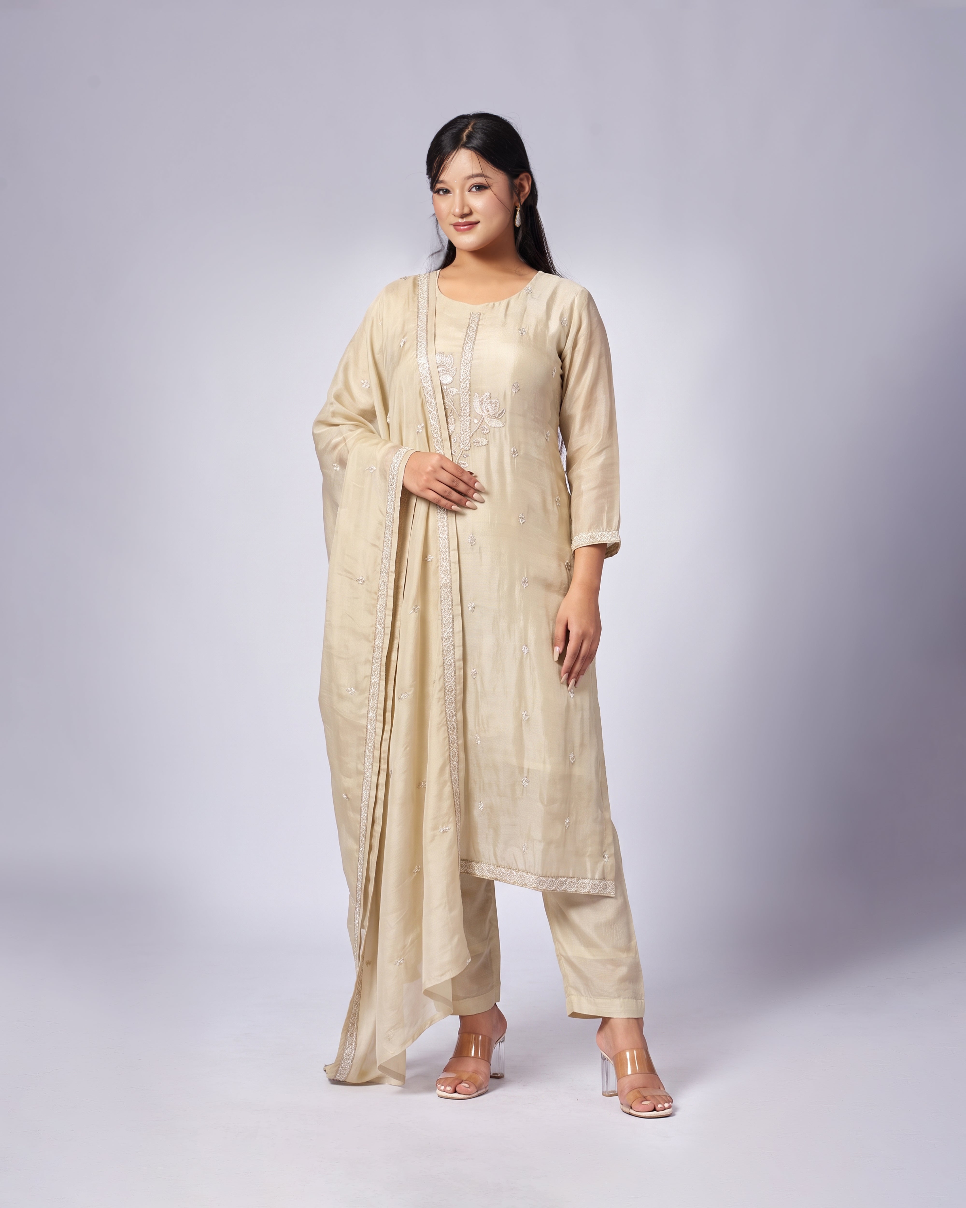 A woman modeling a traditional golden Indian outfit consisting of a kurta, trousers, and a dupatta, with subtle embroidery on the chest, set against a plain light gray studio background.