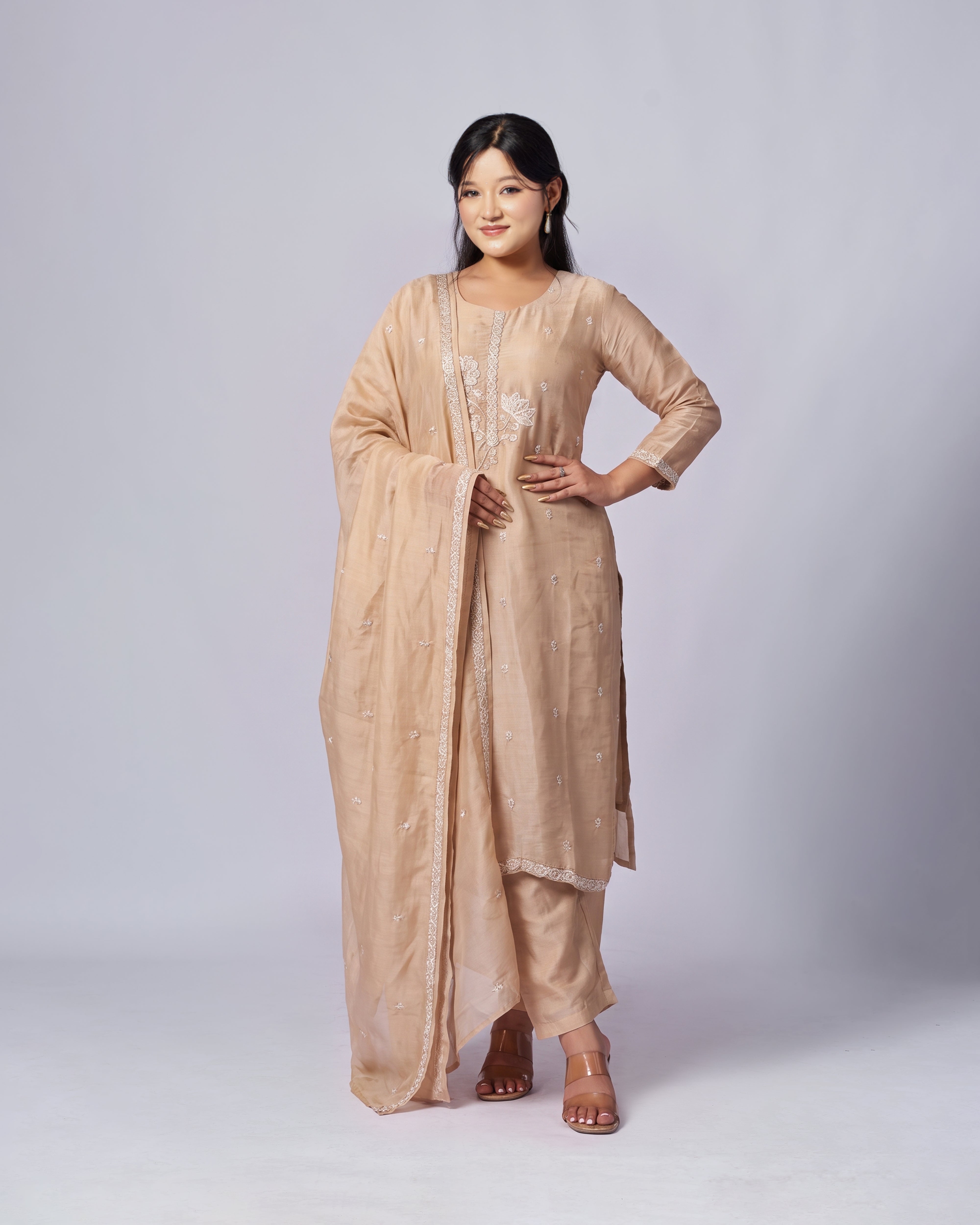 A woman modeling a peach traditional Indian outfit consisting of a kurta, trousers, and a dupatta, with subtle embroidery details, set against a light gray studio background.