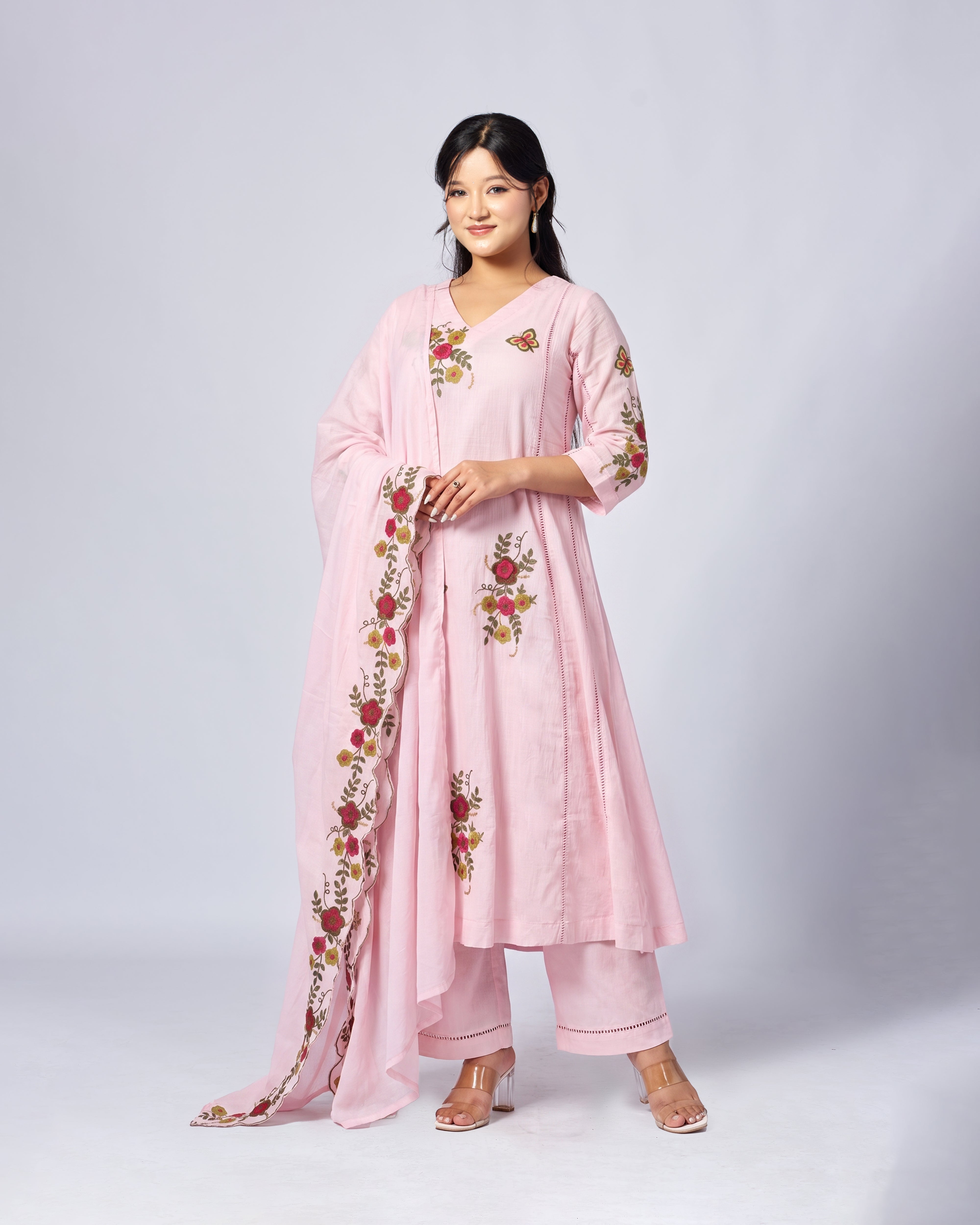 A woman modeling a light pink traditional outfit with matching pants and a draped dupatta, featuring floral embroidery in red, yellow, and green on the kurta, sleeves, and dupatta, set against a light gray studio background.
