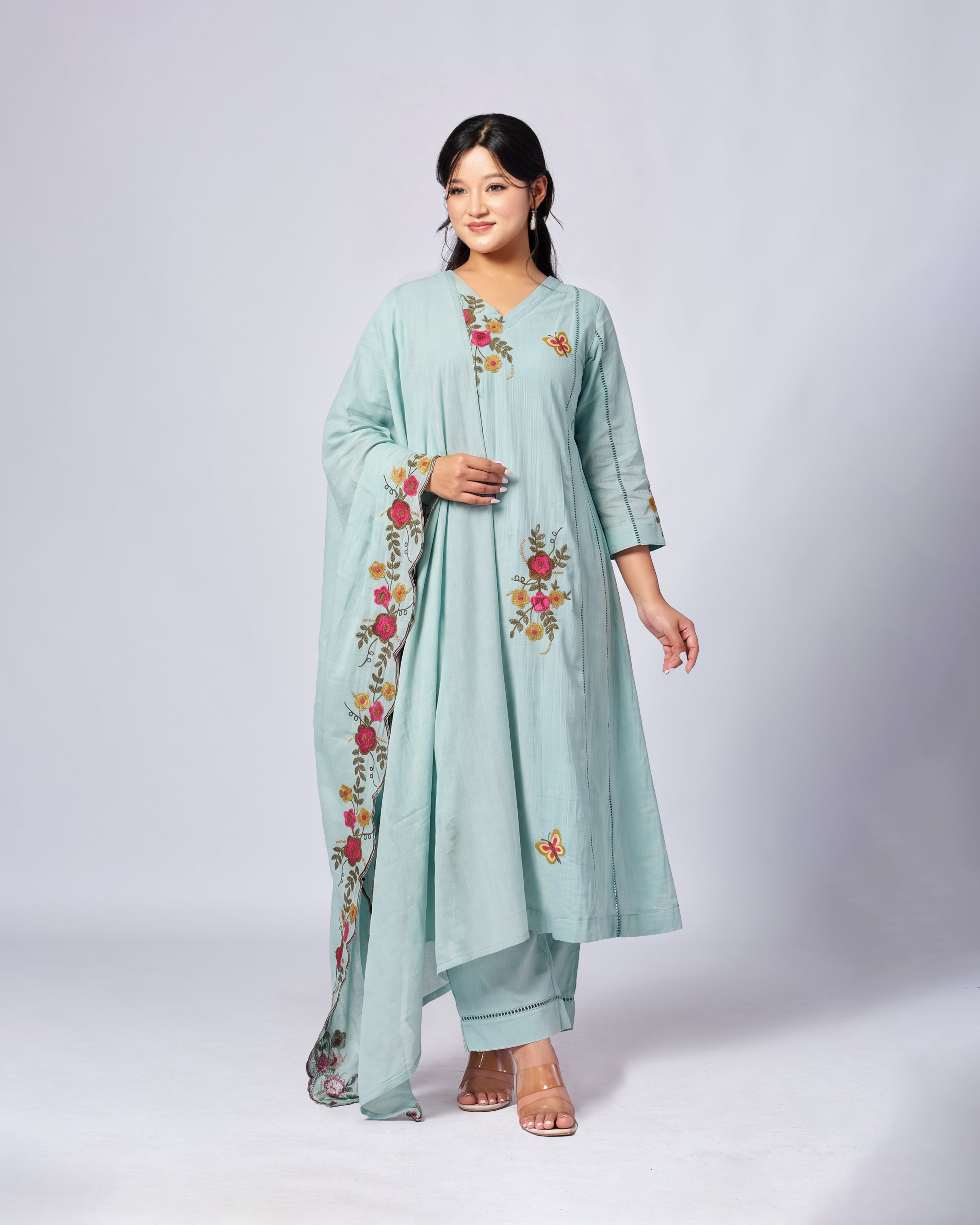 A woman modeling a mint green traditional Indian outfit with floral embroidery and a matching dupatta, set against a plain light gray studio background.