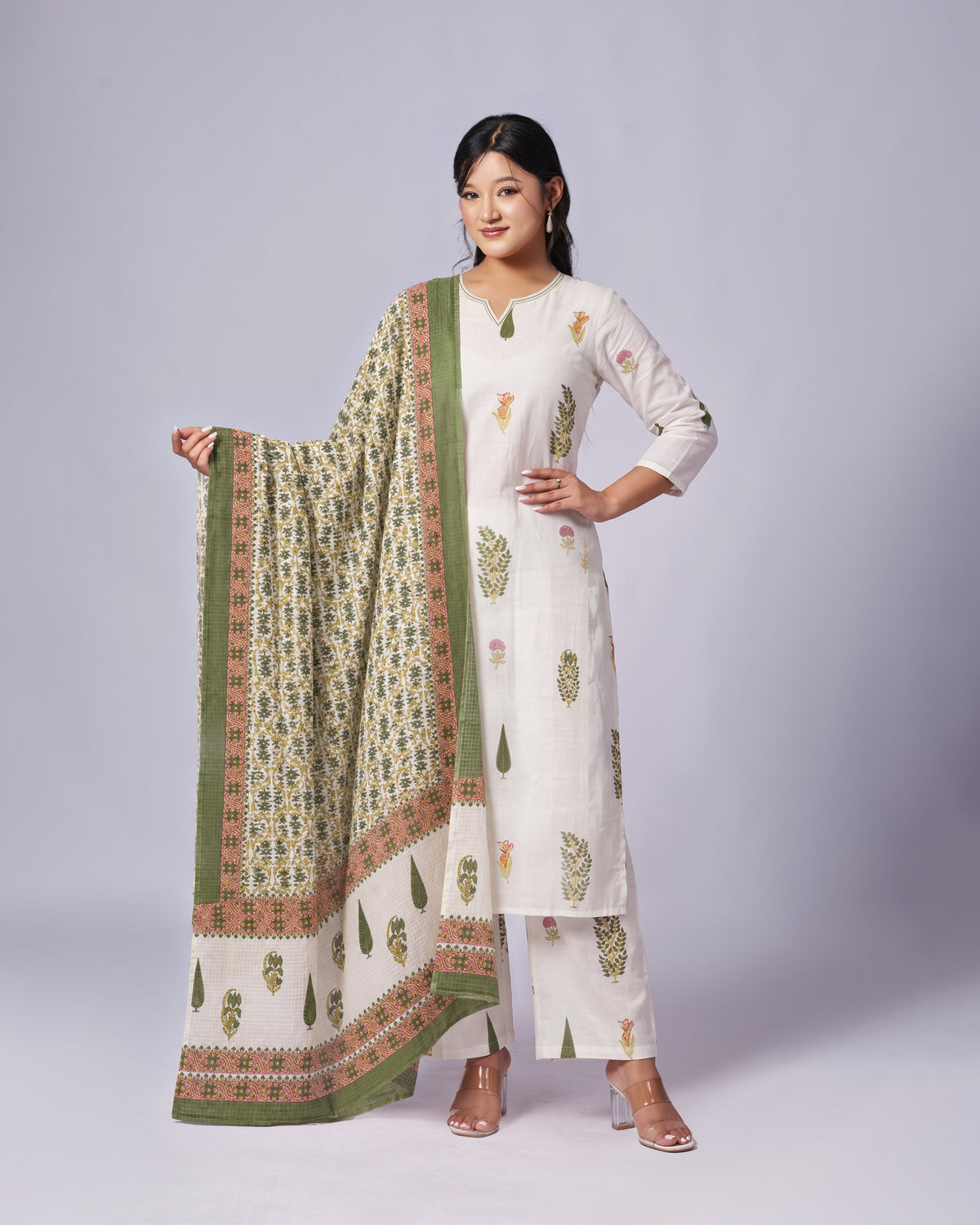 A woman modeling a traditional South Asian salwar kameez set, featuring an off-white kurta and matching trousers with green and gold block prints, paired with a green patterned dupatta, set against a plain light gray studio background.