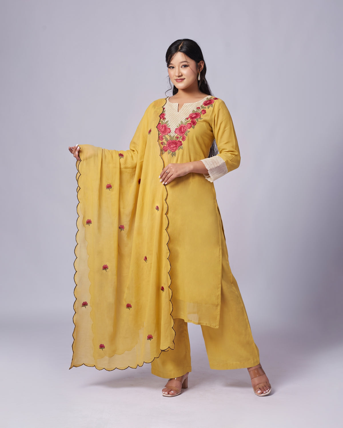 A woman modeling a yellow ethnic salwar kameez suit with a matching dupatta. The kurta features pink floral embroidery around the V-neck and cream-colored sleeve cuffs.