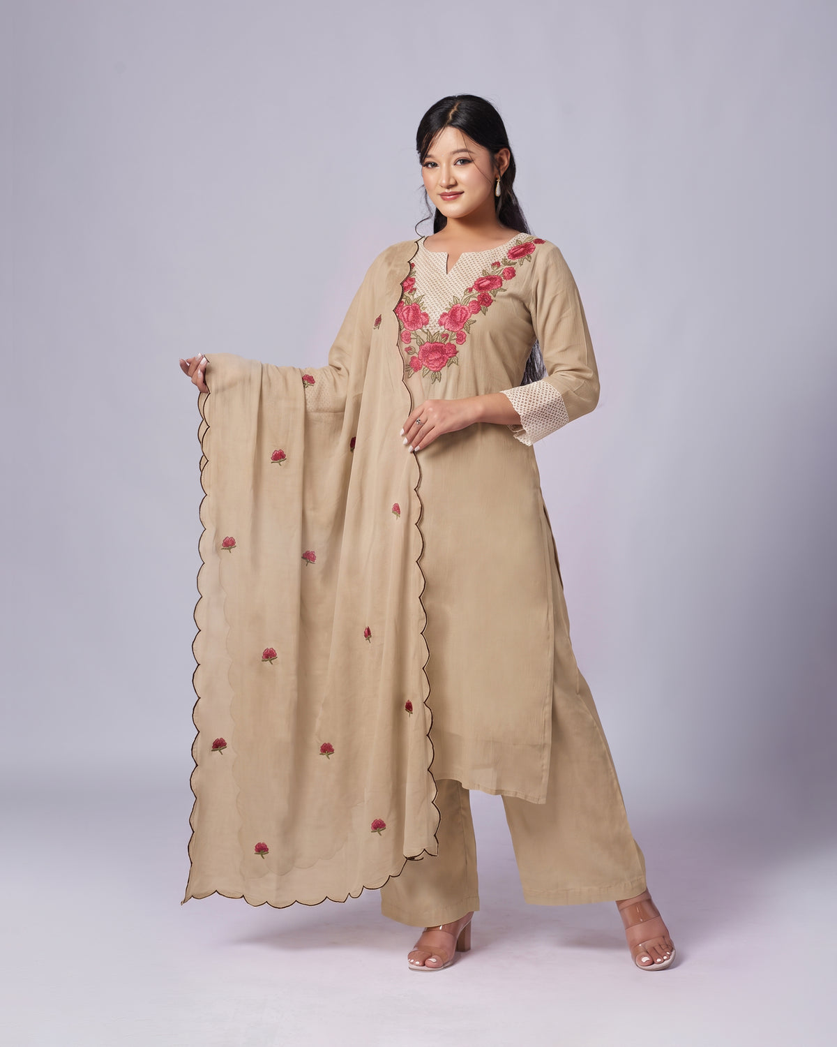 A woman modeling a beige traditional Indian salwar kameez suit with a matching dupatta. The kurta features pink and red floral embroidery on the V-neckline and cuffs, and the dupatta has small matching floral motifs and a scalloped border.
