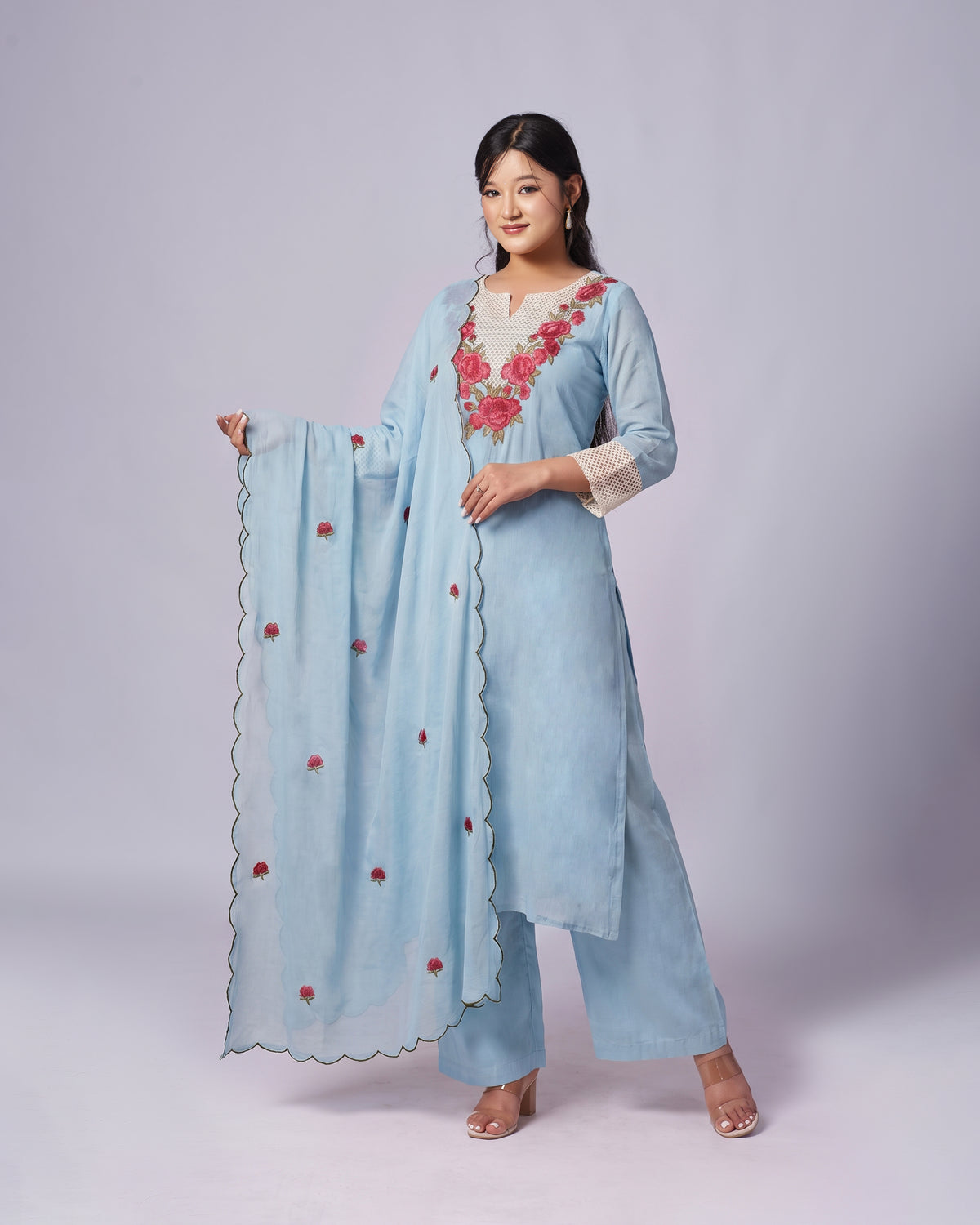 A woman modeling a light blue traditional outfit with a matching dupatta, featuring red floral embroidery on the neckline and scattered along the dupatta border, set against a solid light gray studio background.