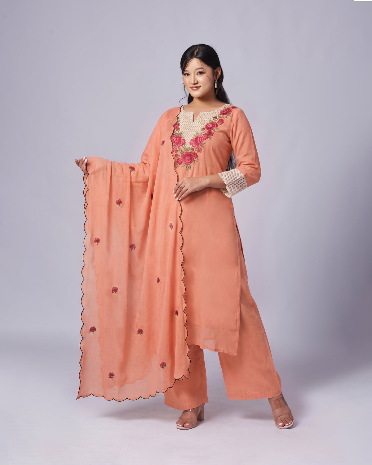 A woman modeling a peach-colored traditional Indian outfit with a matching dupatta, featuring pink floral embroidery on the neckline and scalloped edges.