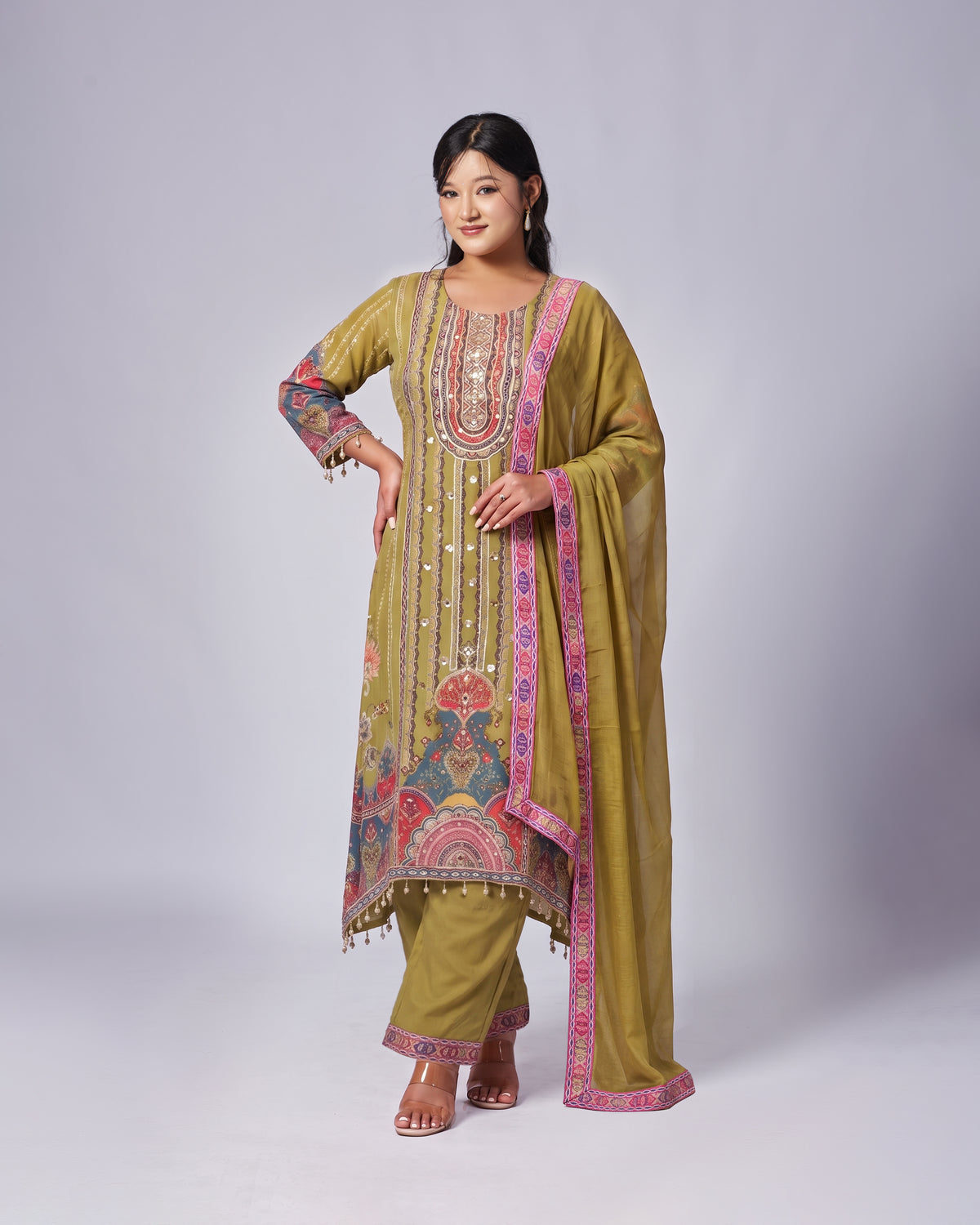 A woman modeling a traditional olive green salwar kameez suit with colorful pink, red, and gold embroidery, paired with matching olive green pants and a sheer dupatta, set against a plain light gray studio background.