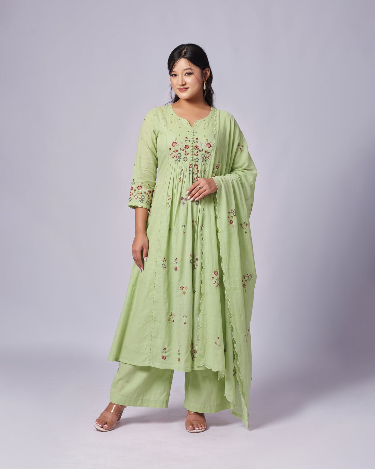 A woman modeling a light green traditional outfit with floral embroidery, consisting of a long kurta, matching pants, and a draped dupatta, set against a plain light gray studio background.