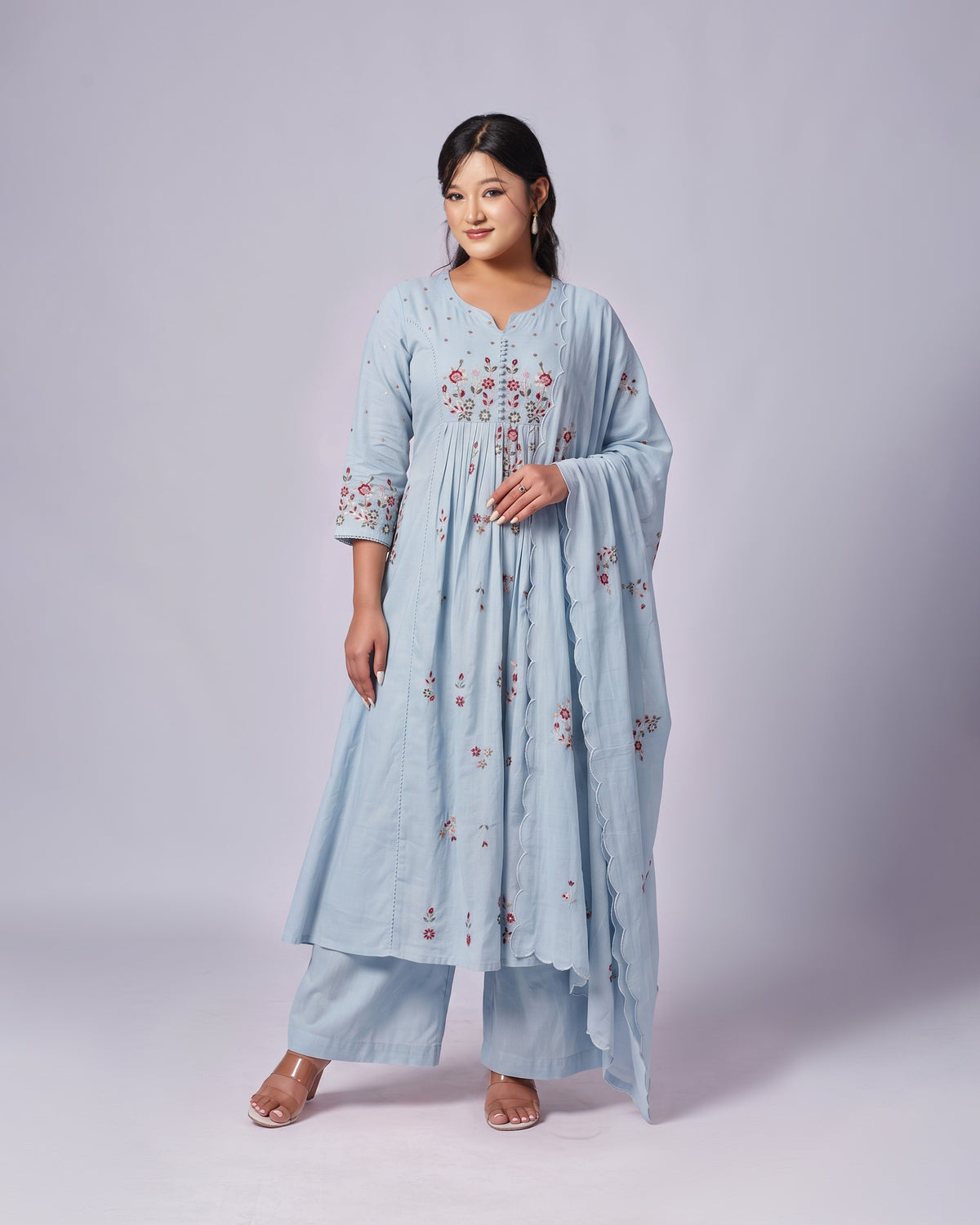A woman modeling a light blue traditional outfit with floral embroidery, consisting of a long kurta, matching pants, and a draped dupatta, set against a plain light gray studio background.