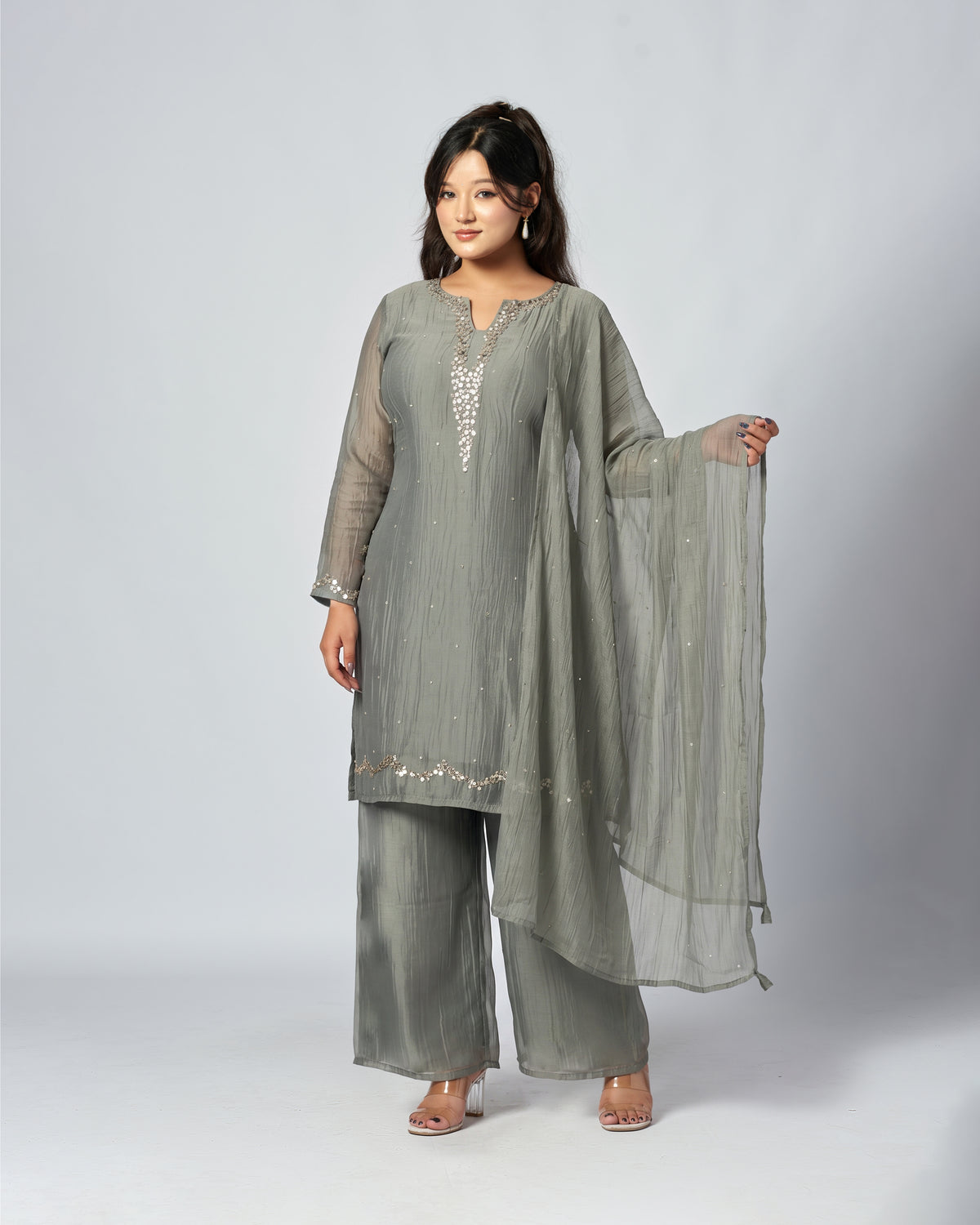 A woman modeling a three-piece traditional outfit in a muted sage green color, featuring a long-sleeved kurta with silver embroidery, matching wide-leg trousers, and a sheer dupatta, set against a plain light gray studio background.