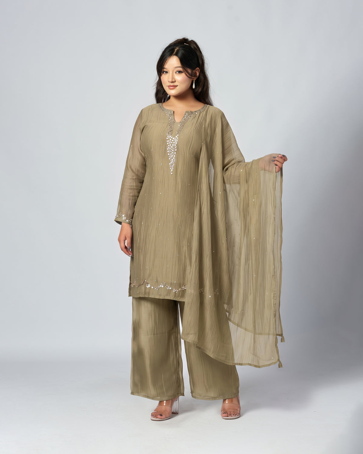 A woman modeling a three-piece traditional outfit in a muted khaki-beige color, featuring a long kurta with silver embellishments, matching wide-leg trousers, and a sheer dupatta, set against a plain light gray studio background.