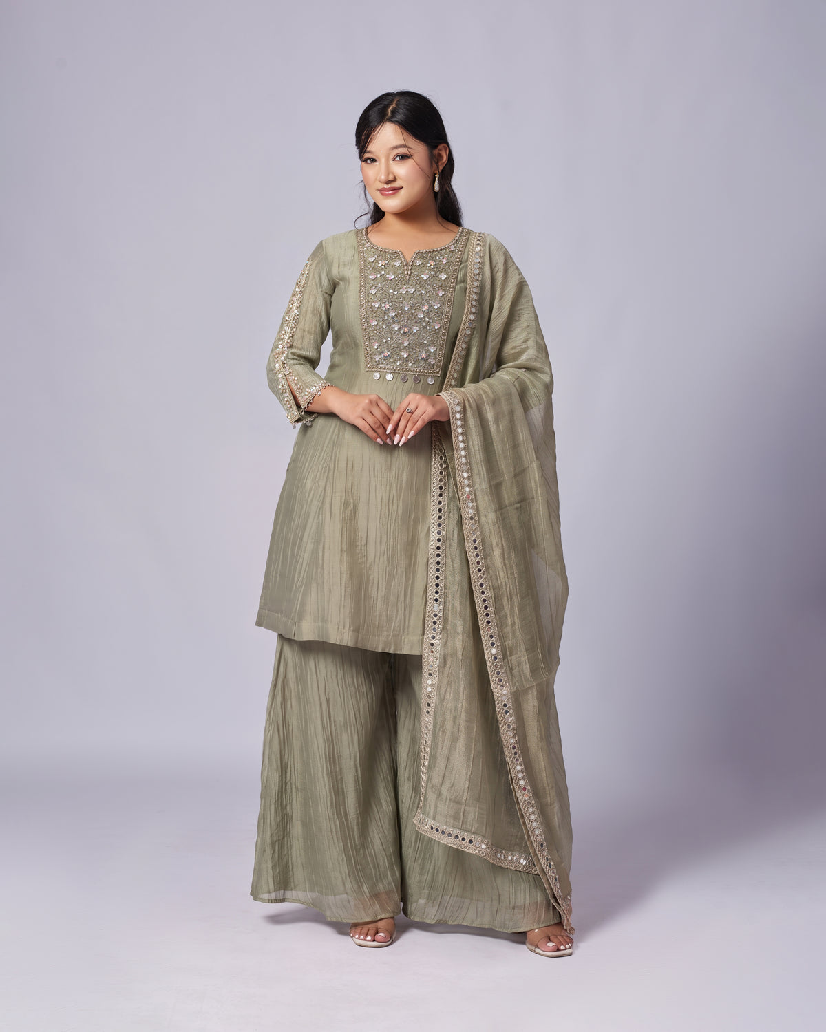A woman modeling a sage green traditional Indian outfit with a matching dupatta, featuring silver mirror work embroidery on the chest, set against a plain light gray studio background.