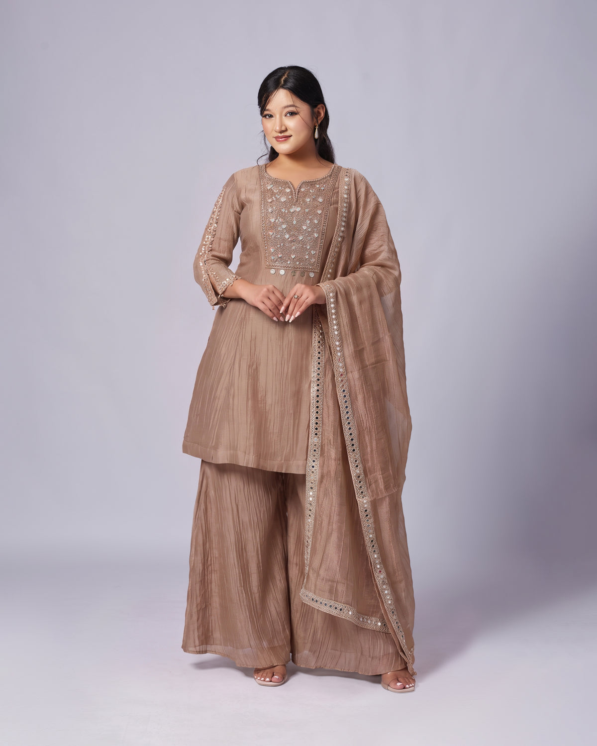 A woman modeling a traditional three-piece salwar kameez suit in a warm taupe-brown color with silver mirror work embroidery on the chest and a matching dupatta.
