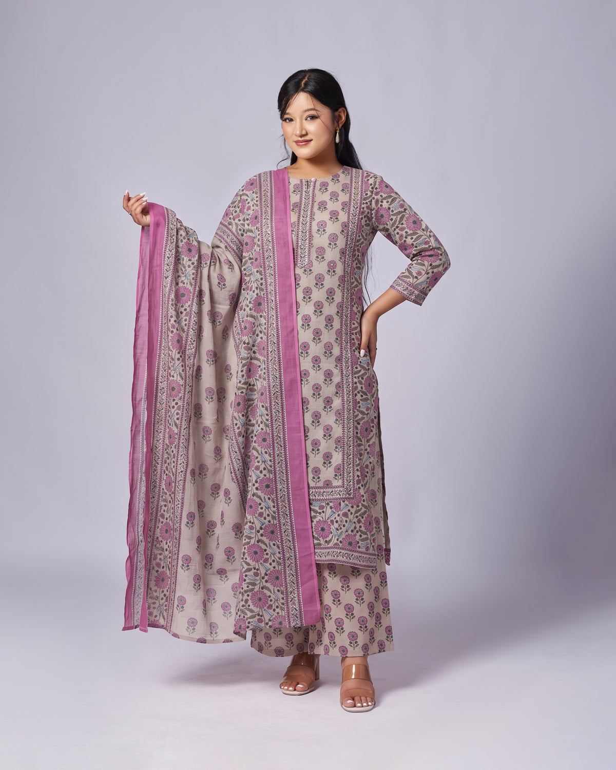 A woman modeling a traditional Indian salwar kameez suit set in a studio setting. The set features a beige kurta and matching pants with pink floral block prints and borders, paired with a matching pink dupatta.