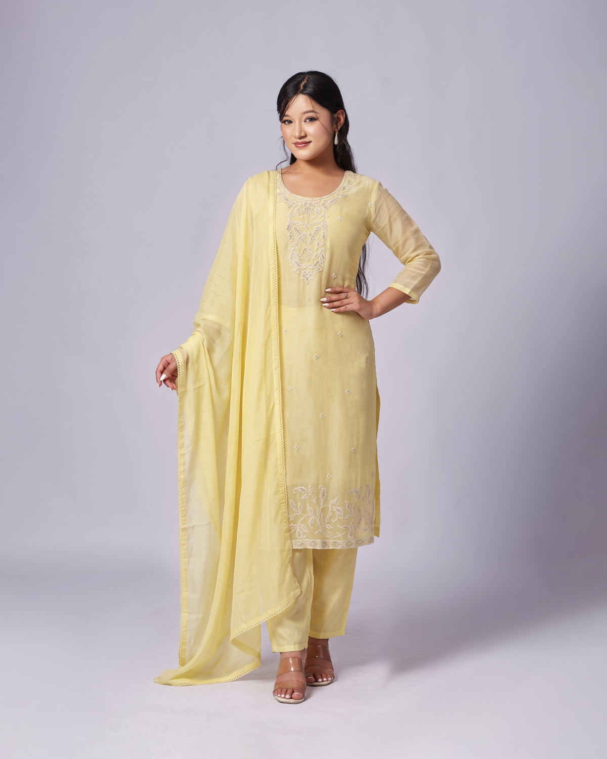 A woman modeling a light yellow ethnic salwar kameez suit with matching dupatta, featuring embroidered details on the kurta and pants, set against a plain light gray studio background.