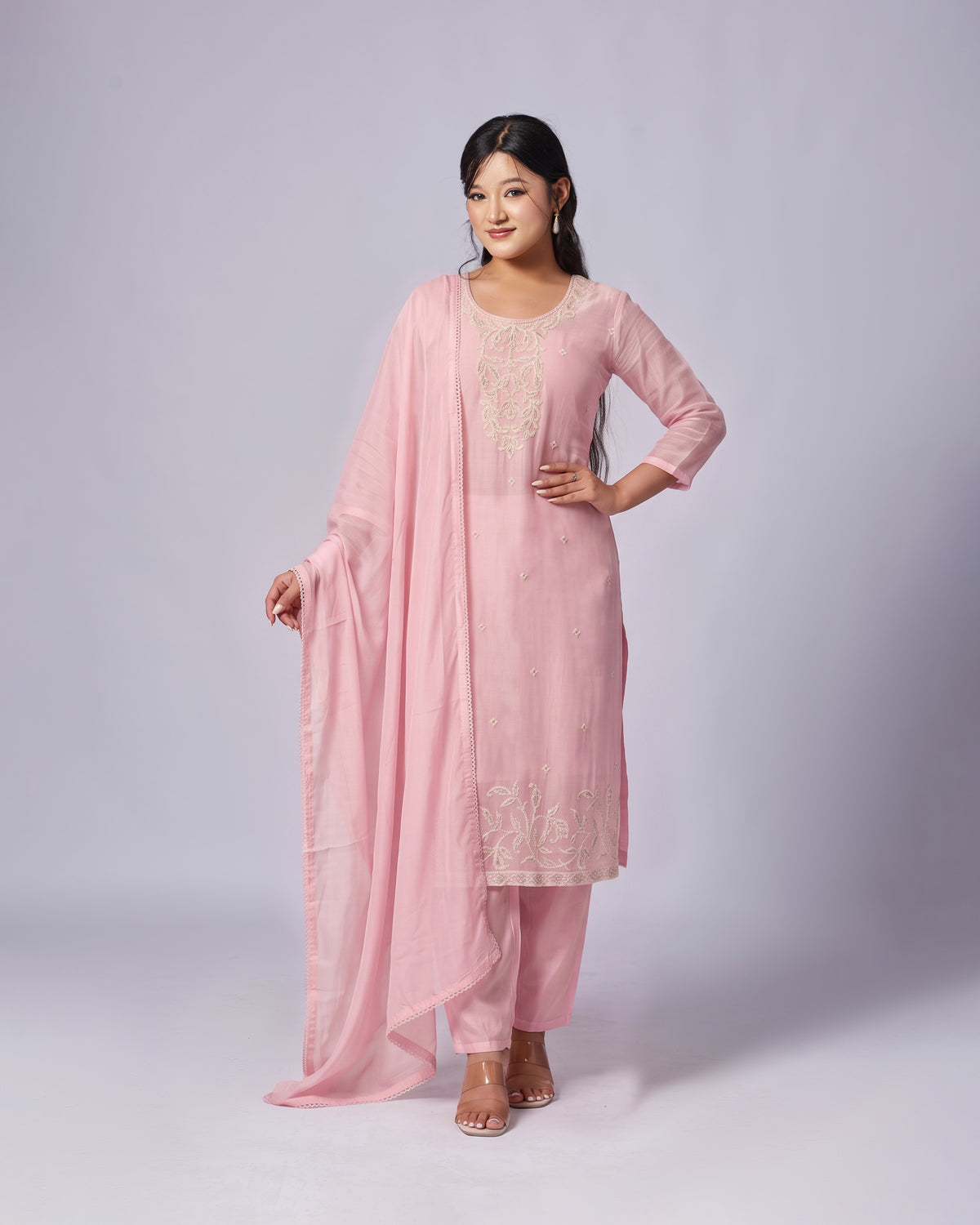 A woman modeling a light pink traditional Indian salwar kameez suit with matching dupatta. The suit features beige floral embroidery on the chest and hem, paired with matching pink pants and clear high-heeled sandals, set against a plain light gray studio background.