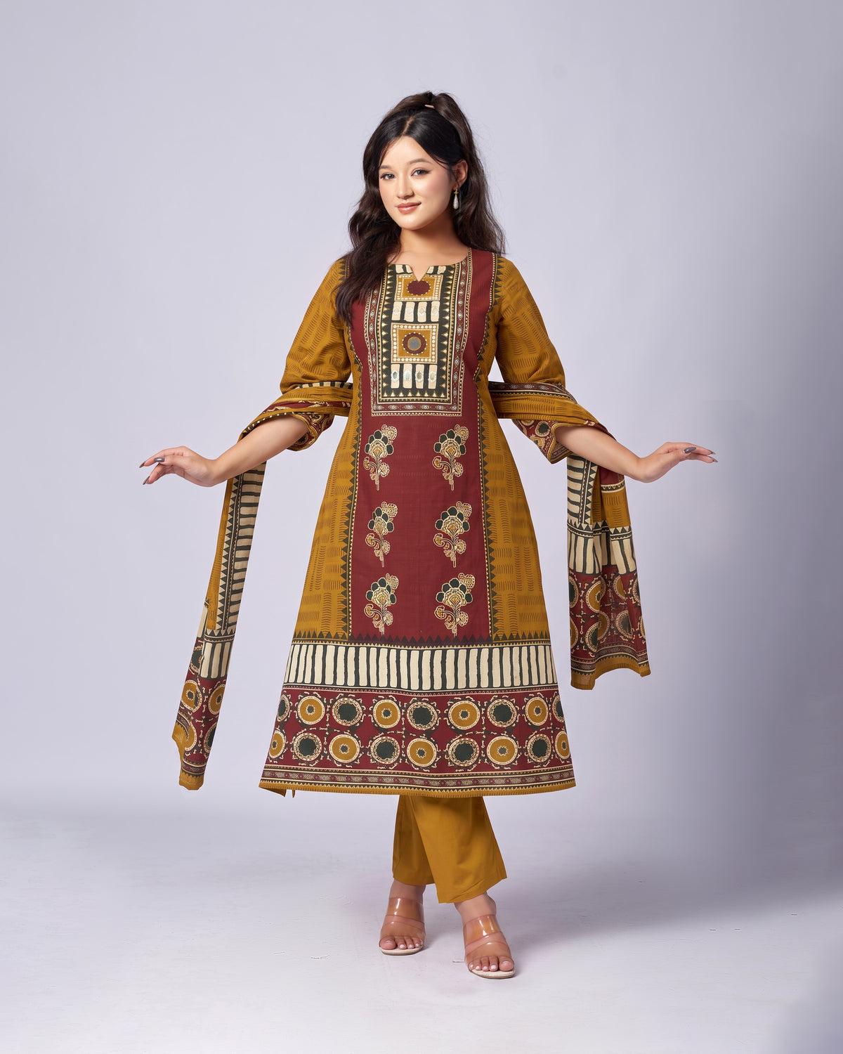 A woman modeling a traditional Indian salwar kameez suit set. The outfit features a mustard yellow kurta with a large maroon front panel decorated with intricate floral and geometric patterns, paired with matching mustard yellow trousers and a dupatta, set against a plain light gray studio background.