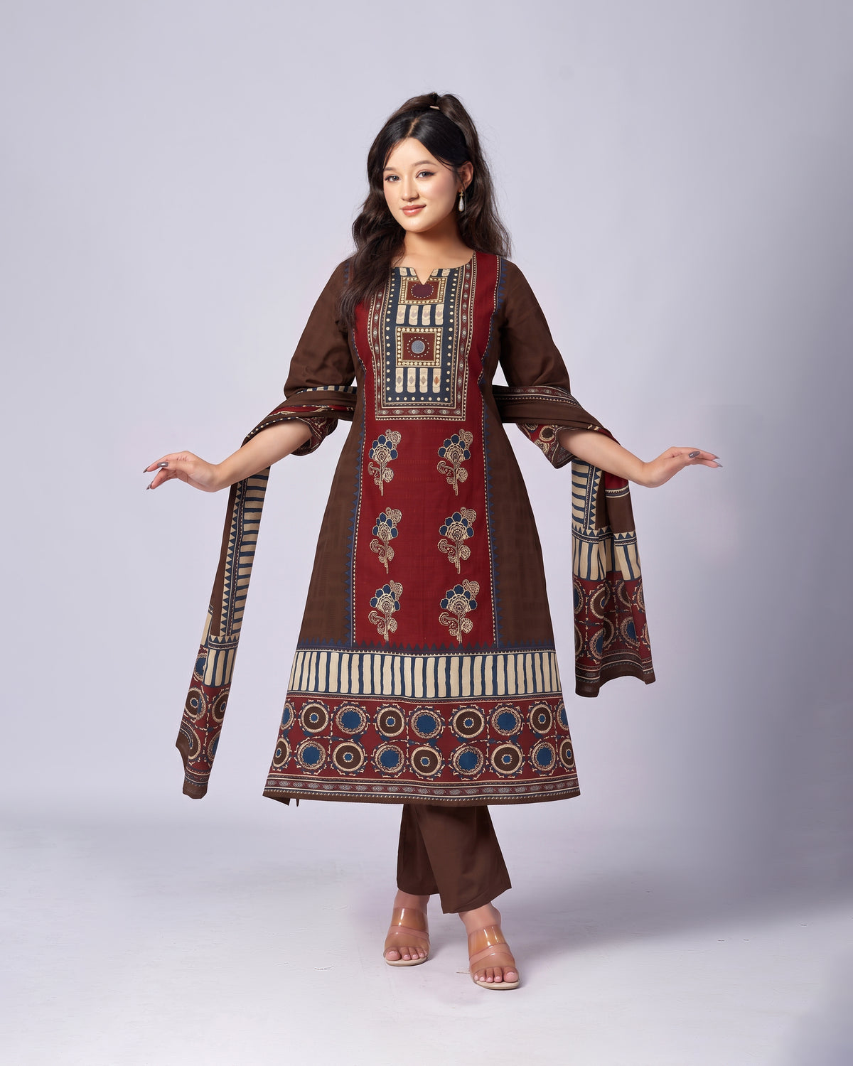 A woman modeling a traditional Indian salwar kameez suit in brown and red with intricate geometric and floral patterns, paired with matching brown trousers, against a light gray studio background.