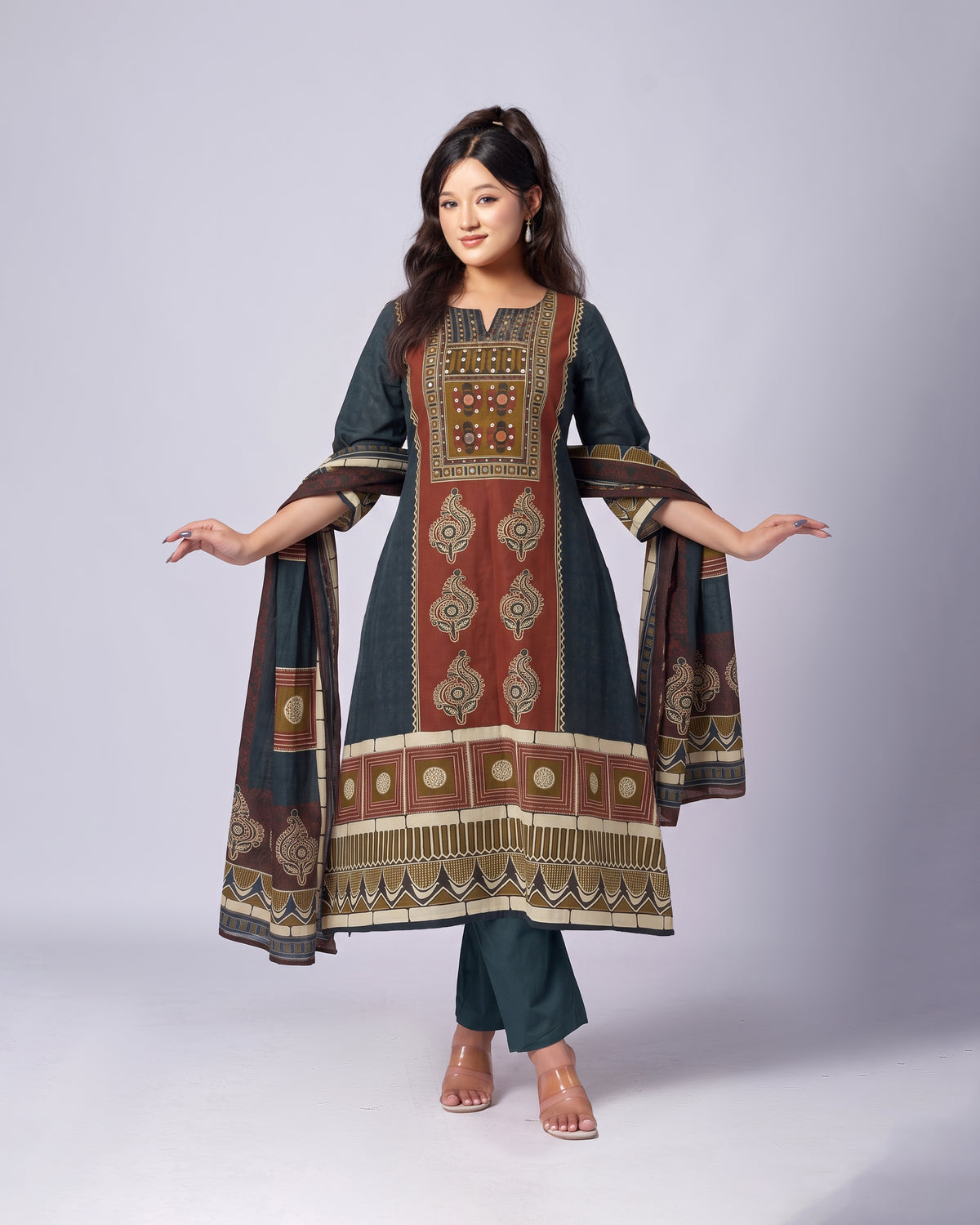 A woman modeling a dark teal ethnic kurta set with a matching dupatta, featuring intricate brown, gold, and beige geometric and floral embroidery, paired with matching dark teal pants, against a light gray studio background.