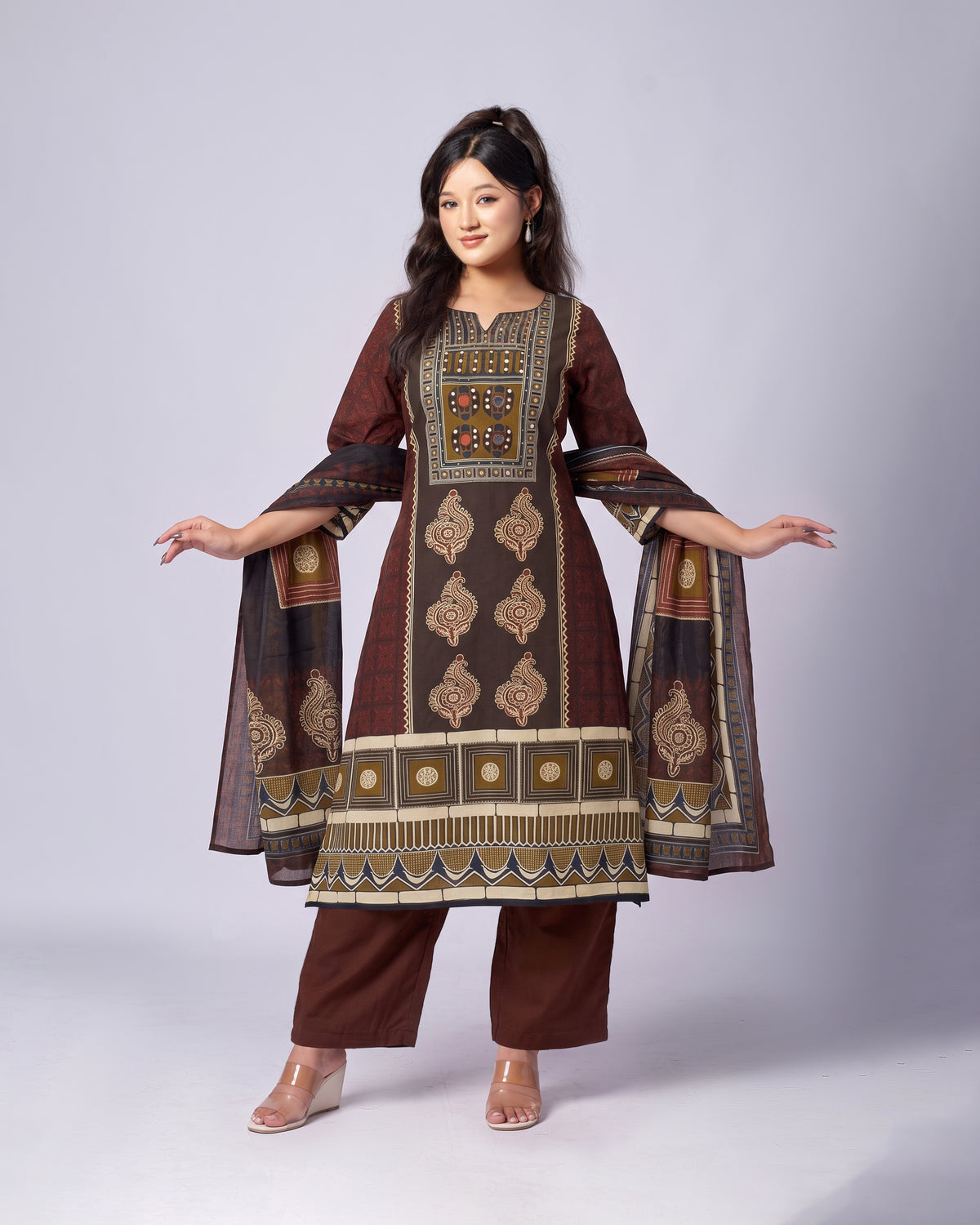 A woman modeling a traditional Indian salwar kameez suit in dark brown with intricate gold, beige, and blue geometric patterns, paired with matching brown trousers and clear wedge sandals, against a light gray studio background.