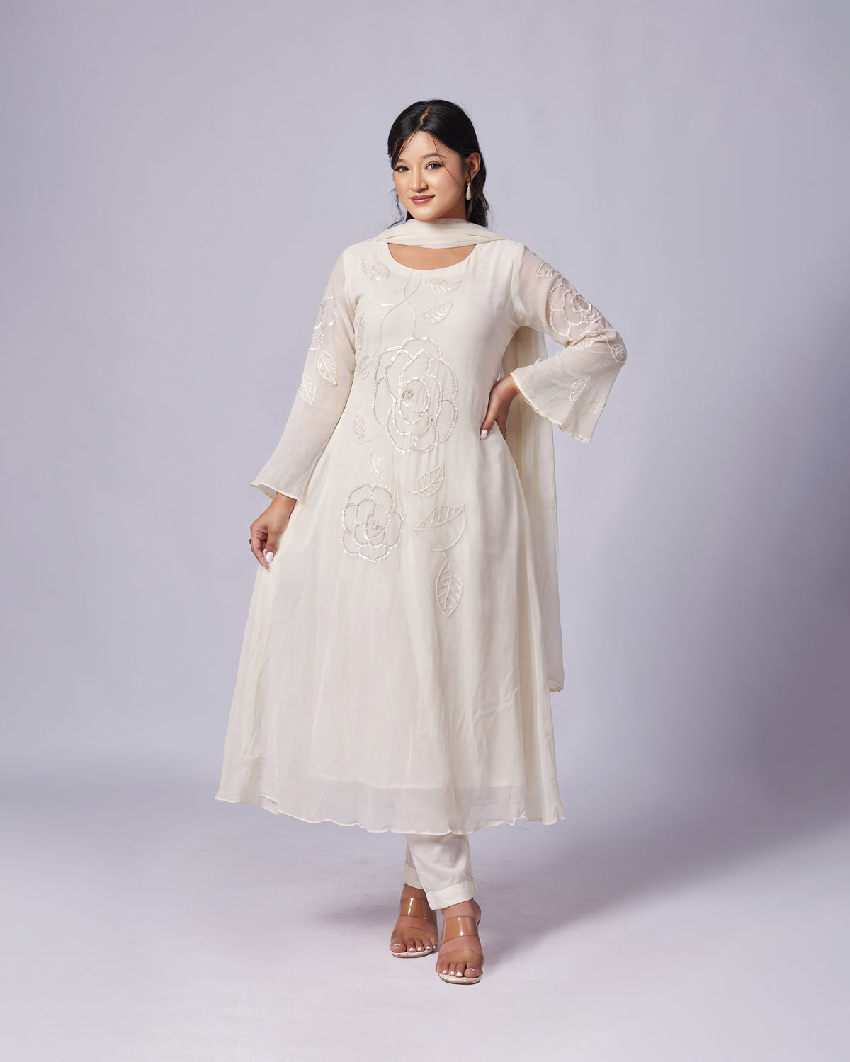 A woman modeling a cream-colored traditional Indian outfit consisting of a long-sleeved embroidered kurta, matching trousers, and a draped dupatta, set against a plain light gray studio background.